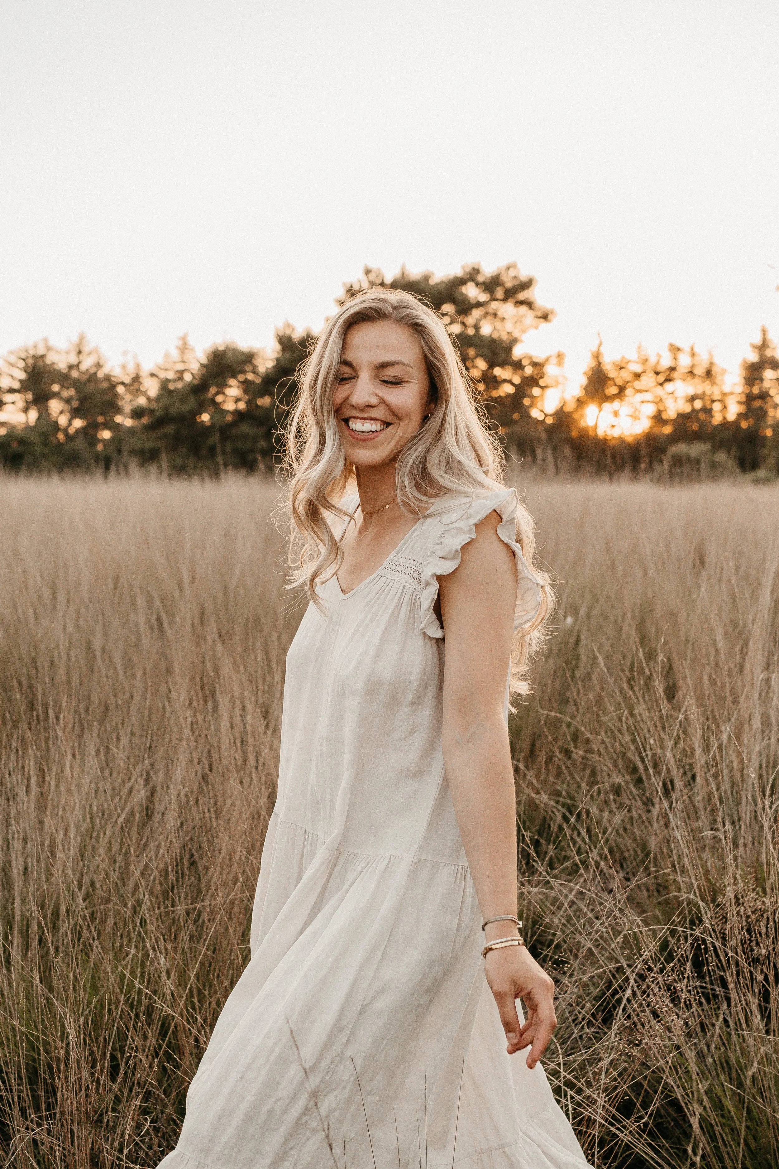 Vrolijke vrouw in een wit jurk die door een veld loopt tijdens zonsondergang, lachend en genietend van de natuur.