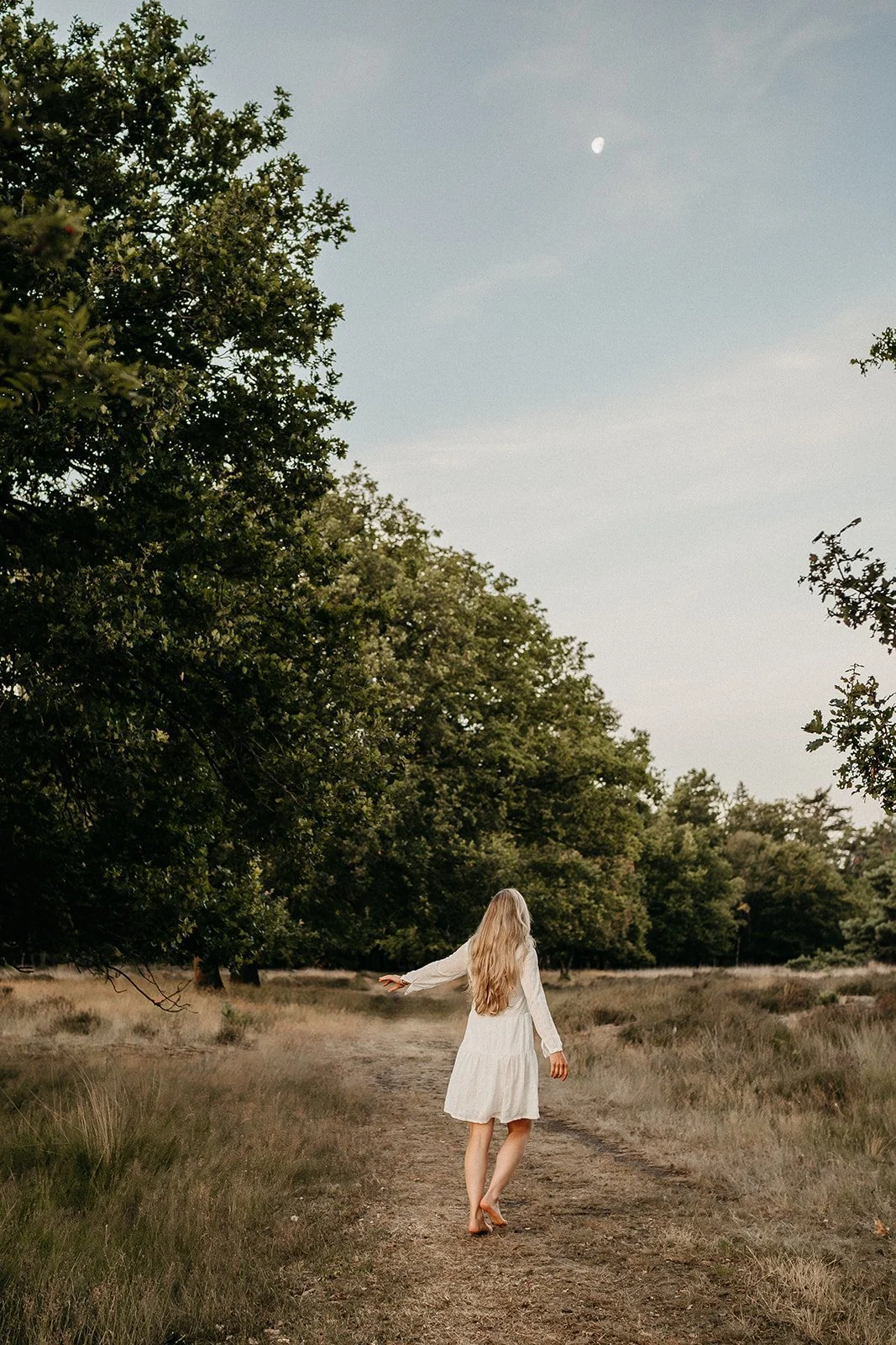 Vrouw in witte jurk wandelt op een pad door een bosrijke omgeving bij zonsondergang of schemering, met een heldere hemel en de maan zichtbaar.