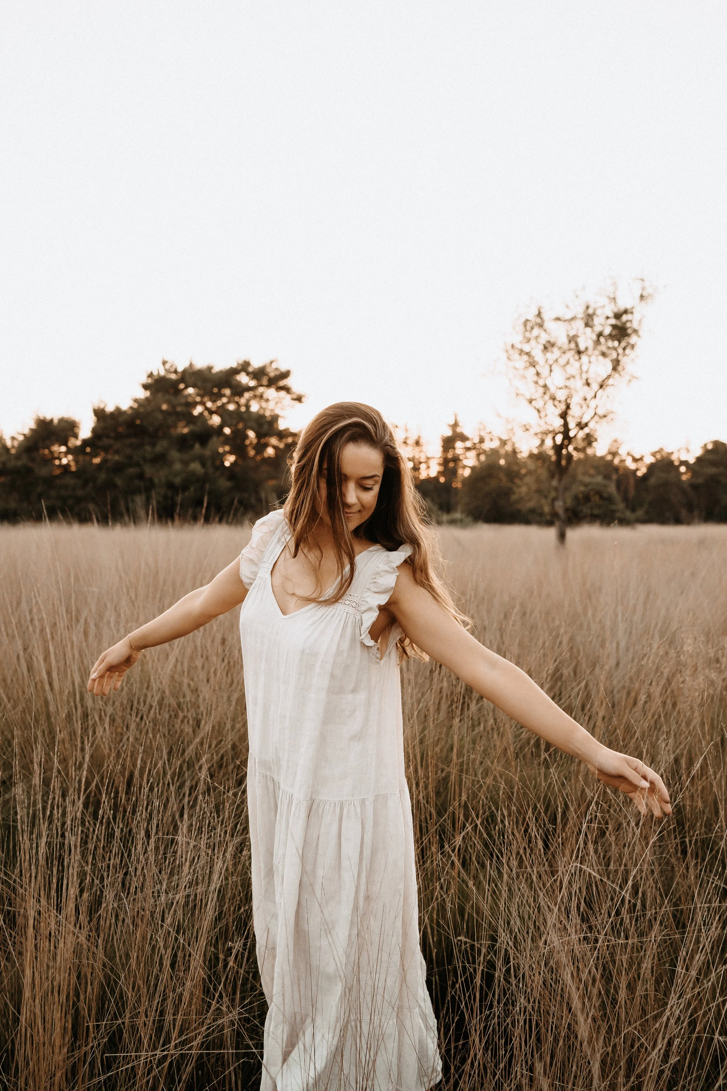 een vrouw in een witte jurk die in een veld met droog gras danst tijdens zonsondergang.