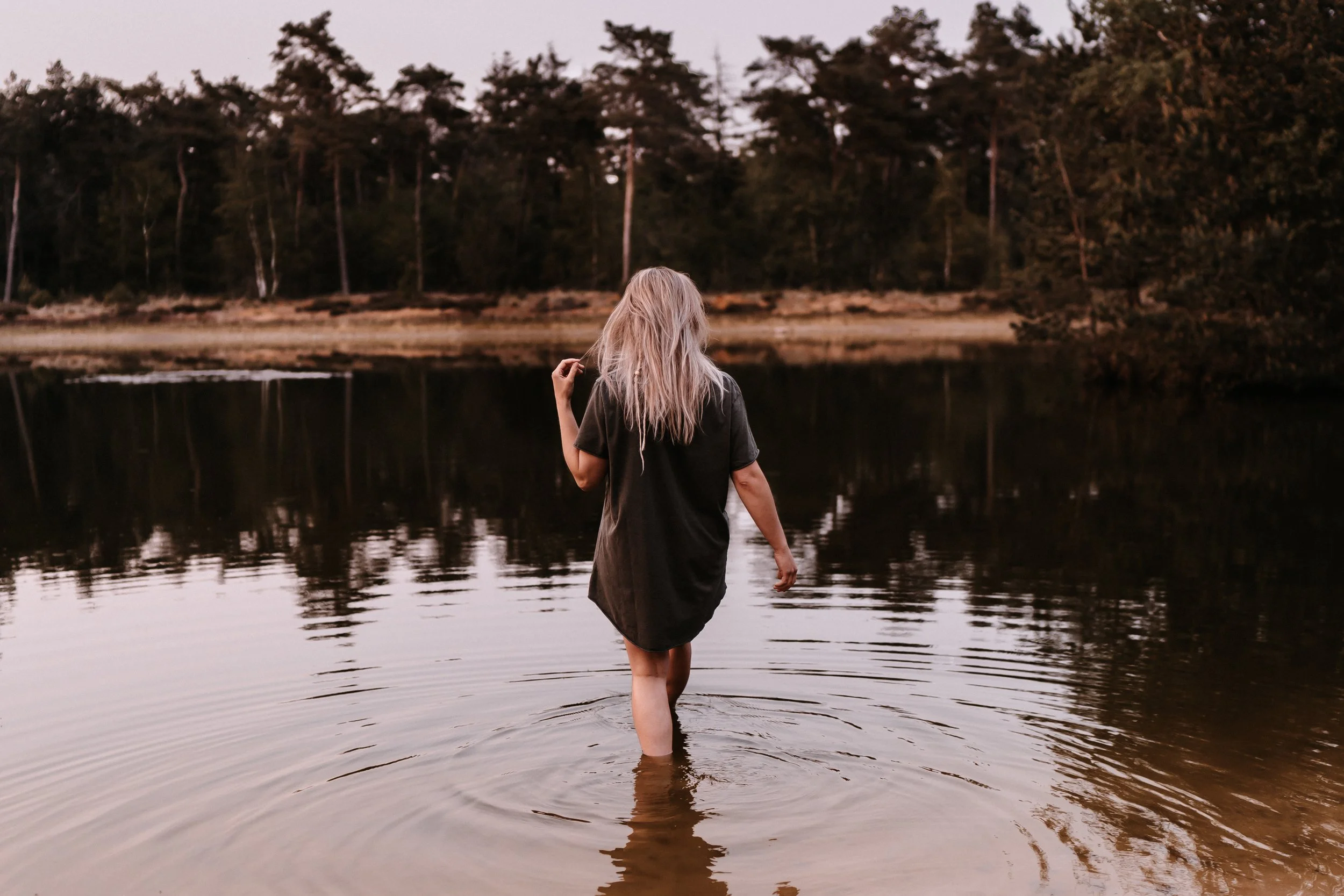 Een vrouw met blond, warrig haar staat half onder water in een rustige rivier of meer, met een bos op de achtergrond, tijdens zonsondergang.