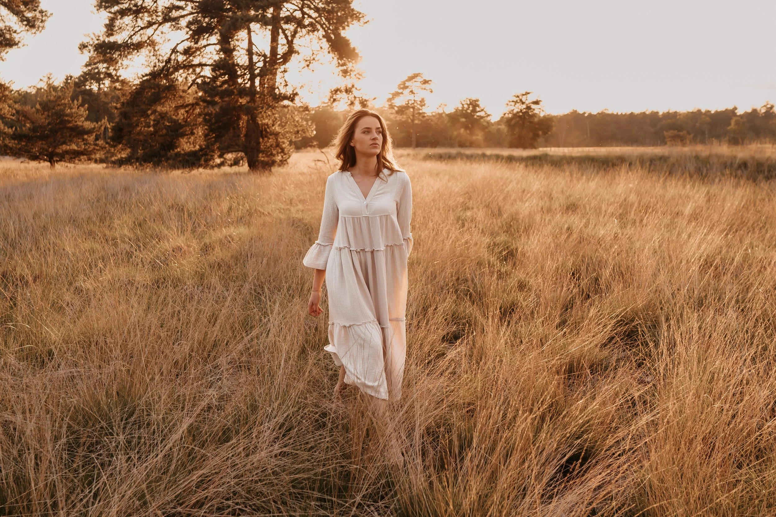 Een vrouw in een witte jurk wandelt door een veld met lang gras tijdens zonsondergang.