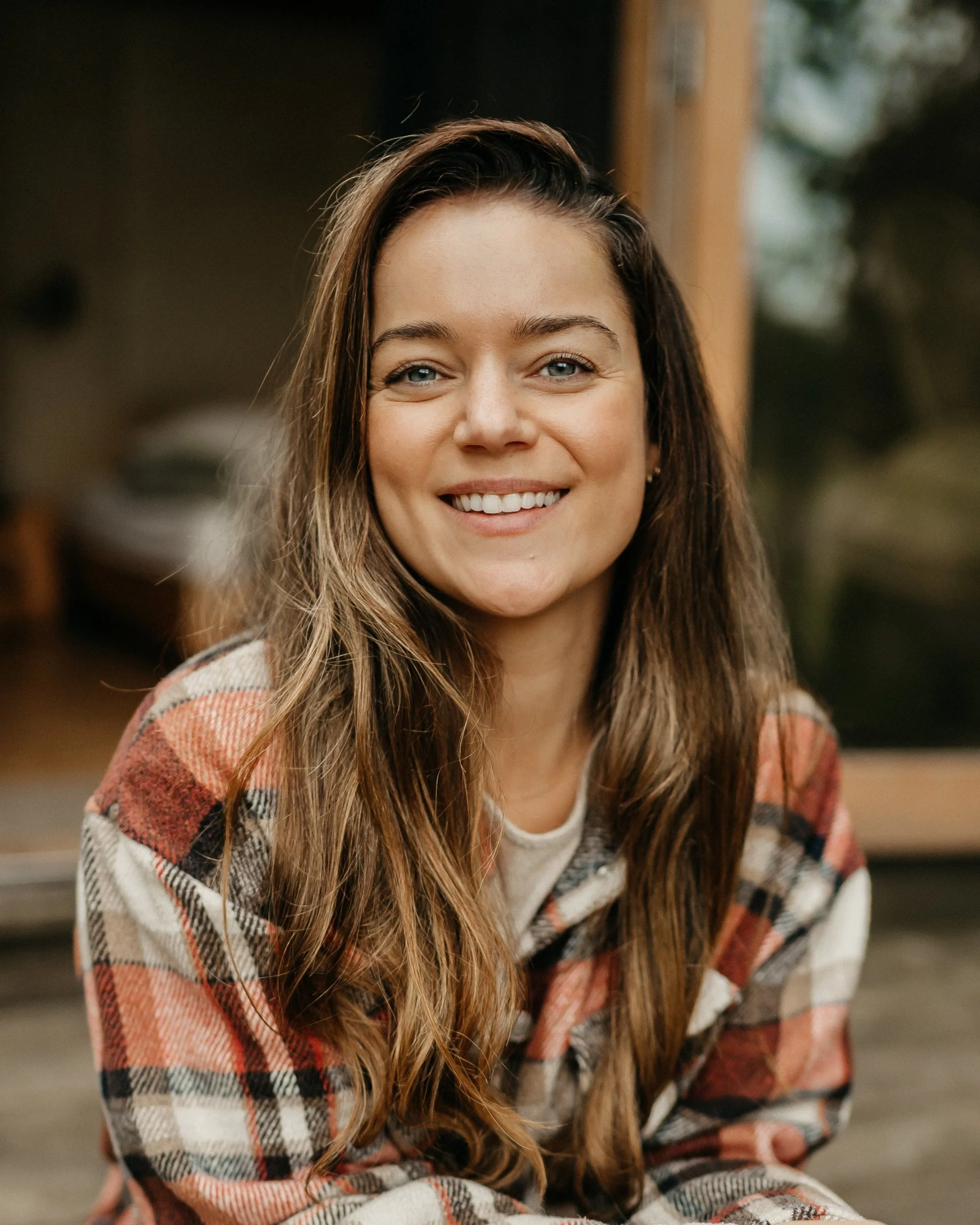 Een glimlachende vrouw met lange, golvende bruintoene haar, gekleed in een rood-wit-zwart geruit overhemd, zittend bij een raam met een natuurlijke achtergrond.