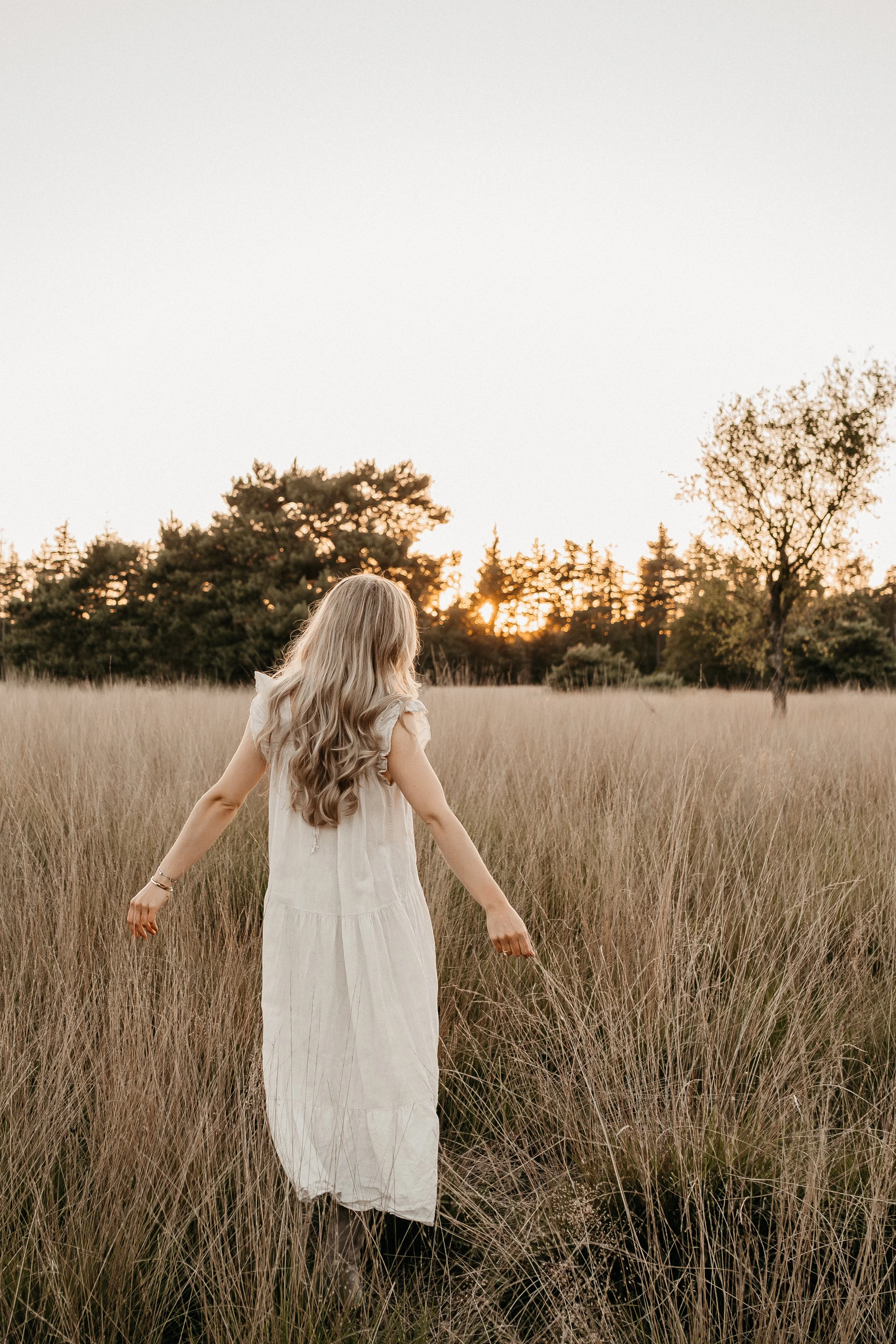 Een vrouw met lange, golvende blonde haar die door een grasveld loopt tijdens zonsondergang.