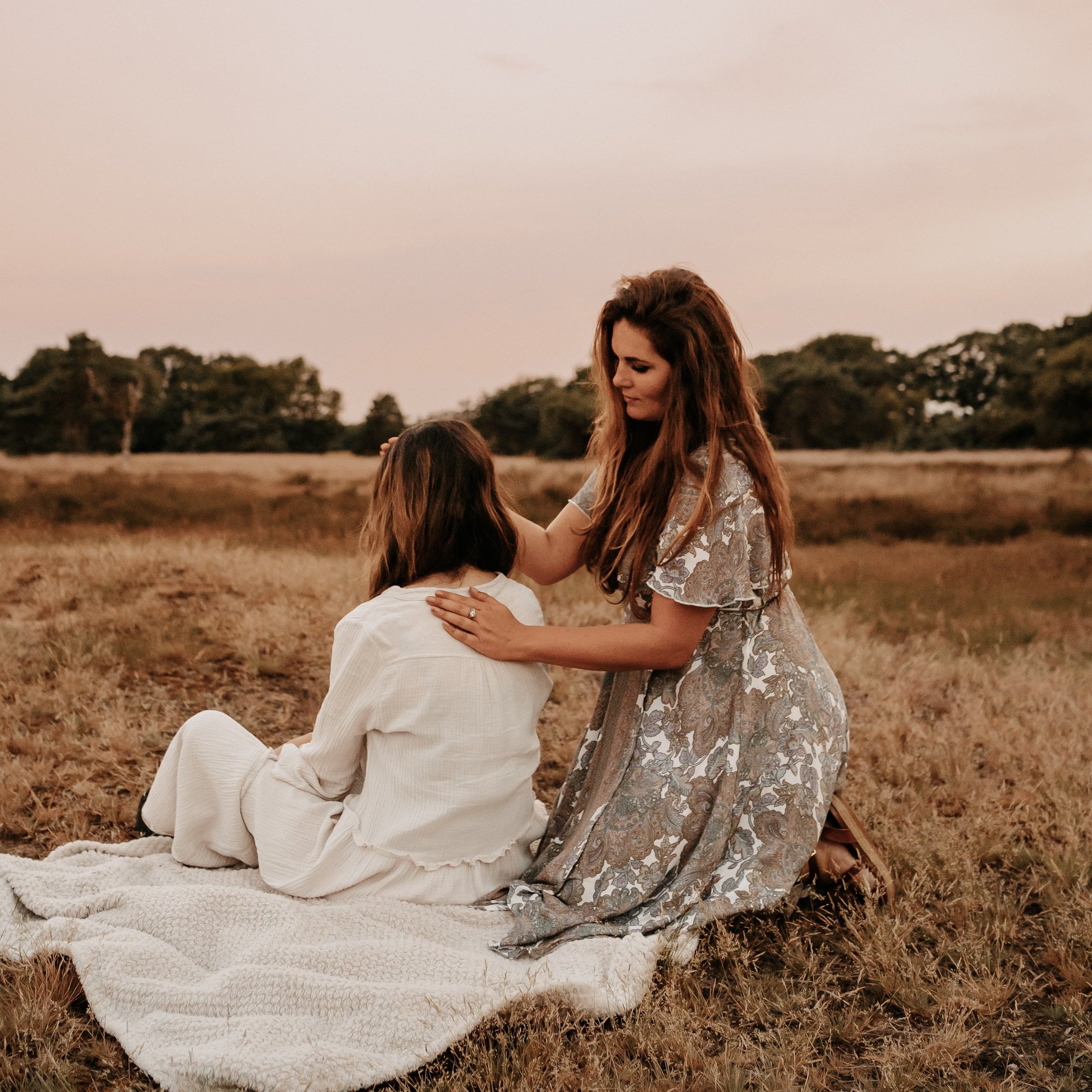 Twee vrouwen op een veld tijdens zonsondergang, een vrouw zit op een witte deken en de ander knielt naast haar, ze omhelzen elkaar liefdevol.