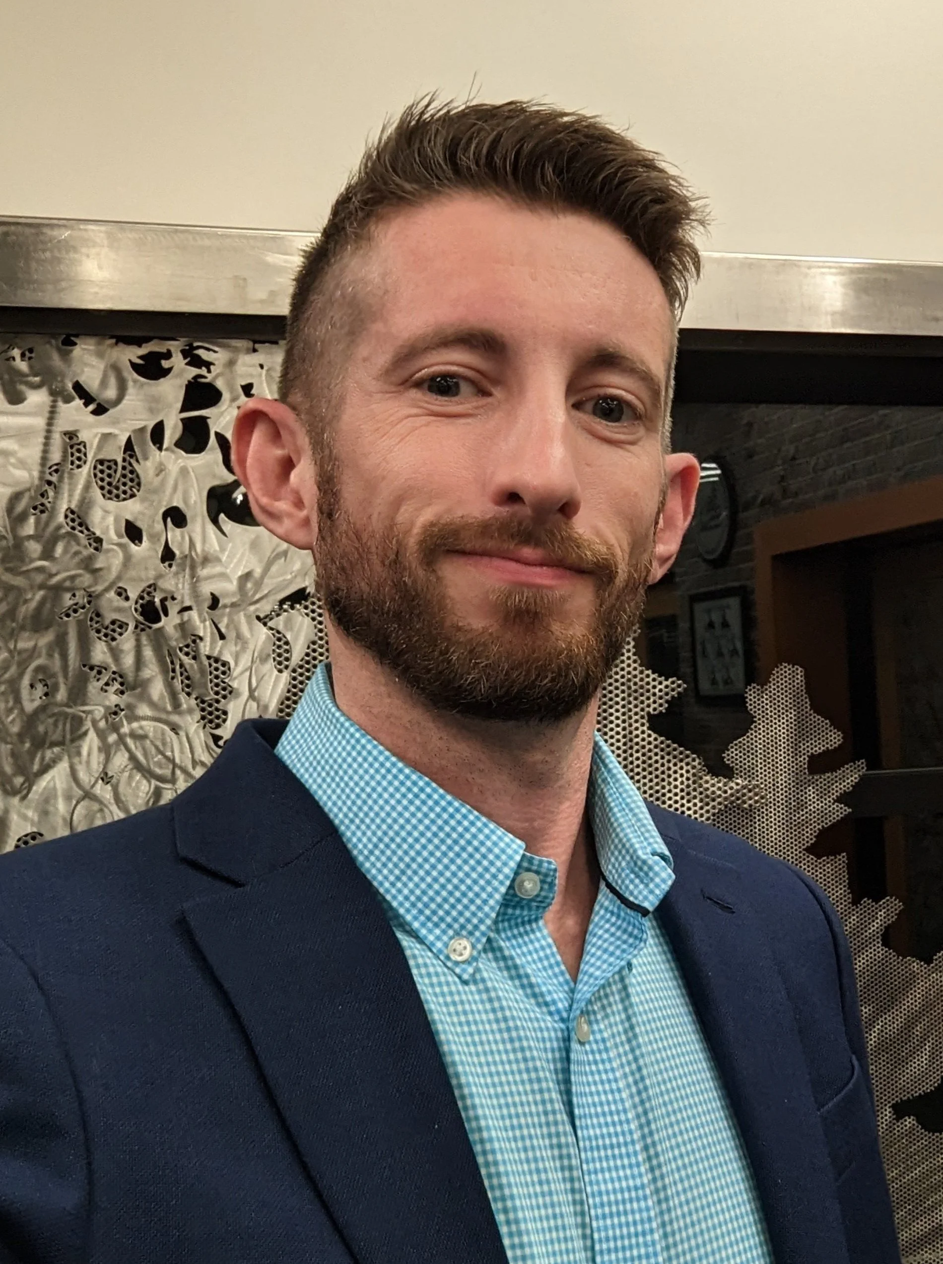 A man with short brown hair, a trimmed beard and mustache, wearing a navy blazer and a blue checkered shirt, smiling at the camera in an indoor setting.