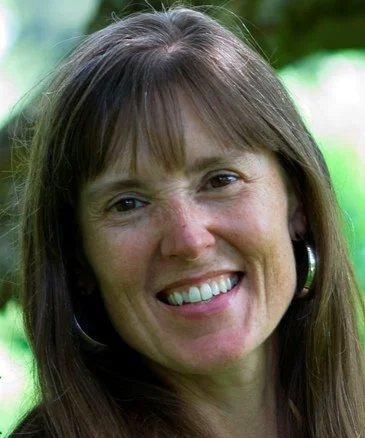 A picture of Karen Story, She has long, straight brown hair with bangs and is wearing large hoop earrings. The photo is taken outdoors with soft, natural light and a background of green foliage.