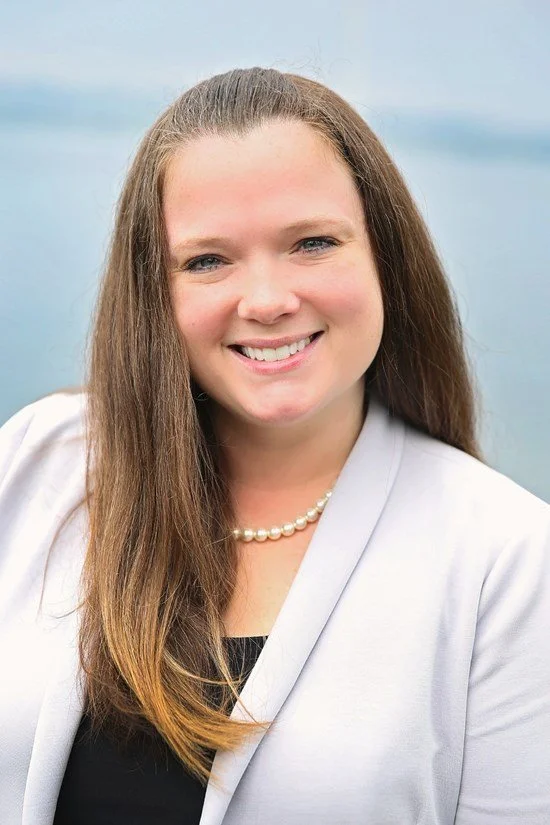 Portrait of a young woman with long brown hair, smiling, wearing a light-colored blazer, black top, and pearl necklace, outdoors with a blue sky background.