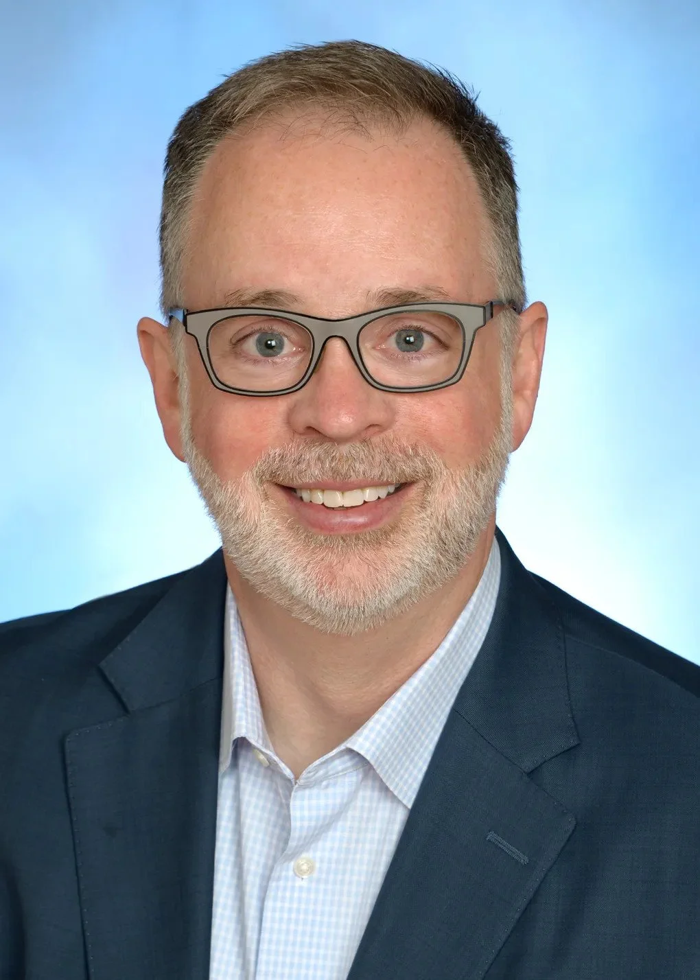 A smiling man with glasses, a beard, and short hair, wearing a dark suit and light-colored shirt against a soft blue background.