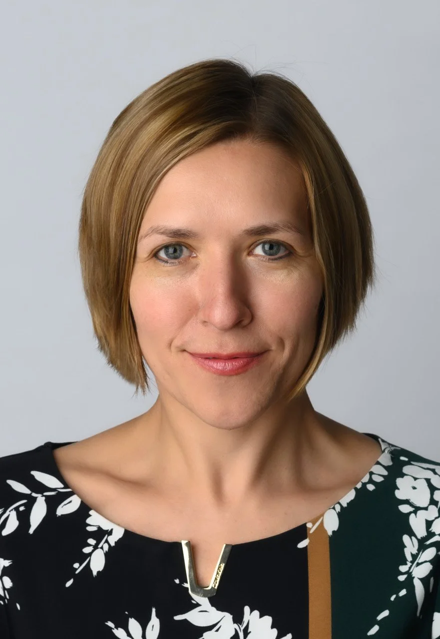 A woman with short, light brown hair and blue eyes smiling at the camera, wearing a black top with white floral patterns and a metal accent on the neckline.