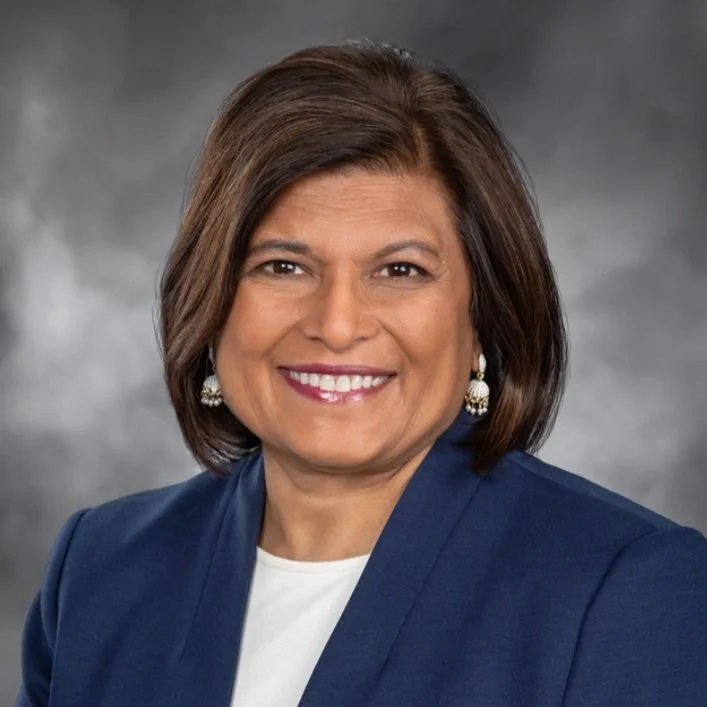 Professional woman with shoulder-length brown hair, wearing a navy blazer and pearl earrings, smiling in a studio portrait with a gray background.