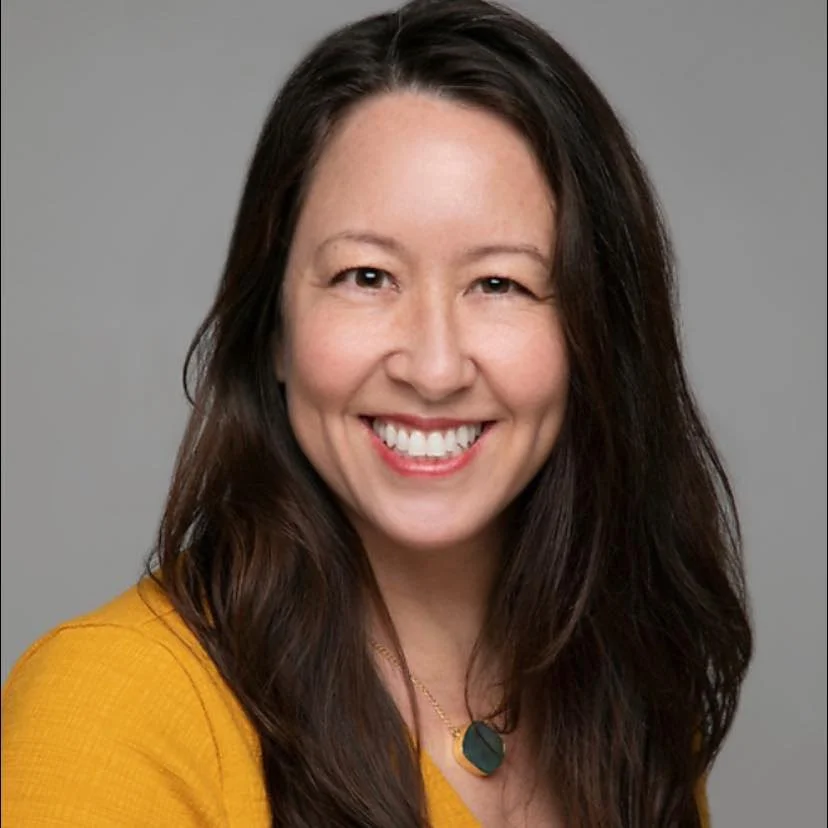 A woman with long dark hair smiling, wearing a mustard yellow top and a green pendant necklace, against a gray background.