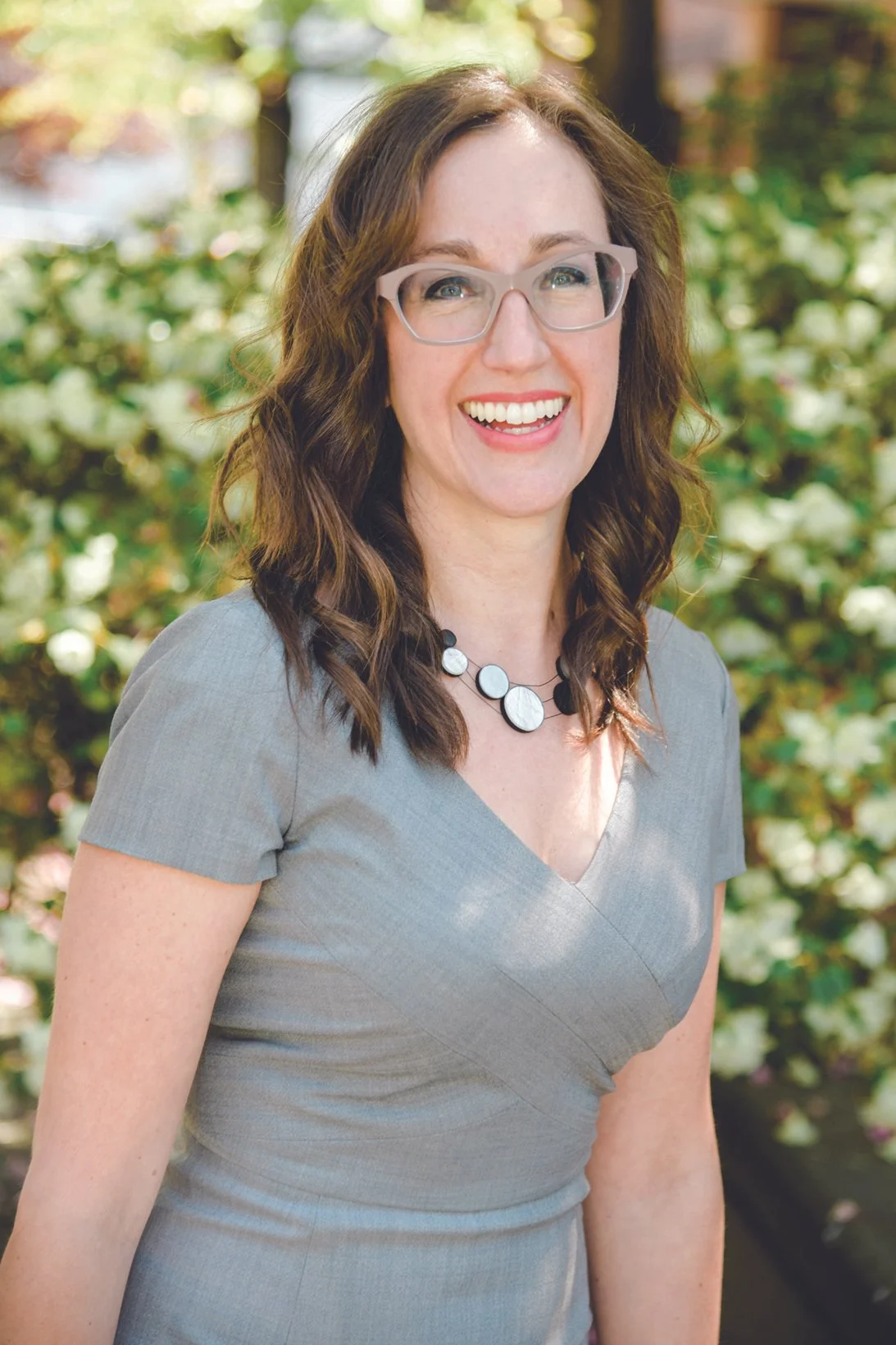 A woman with brown wavy hair, wearing glasses and a gray dress, smiling outdoors with greenery and white flowers in the background.
