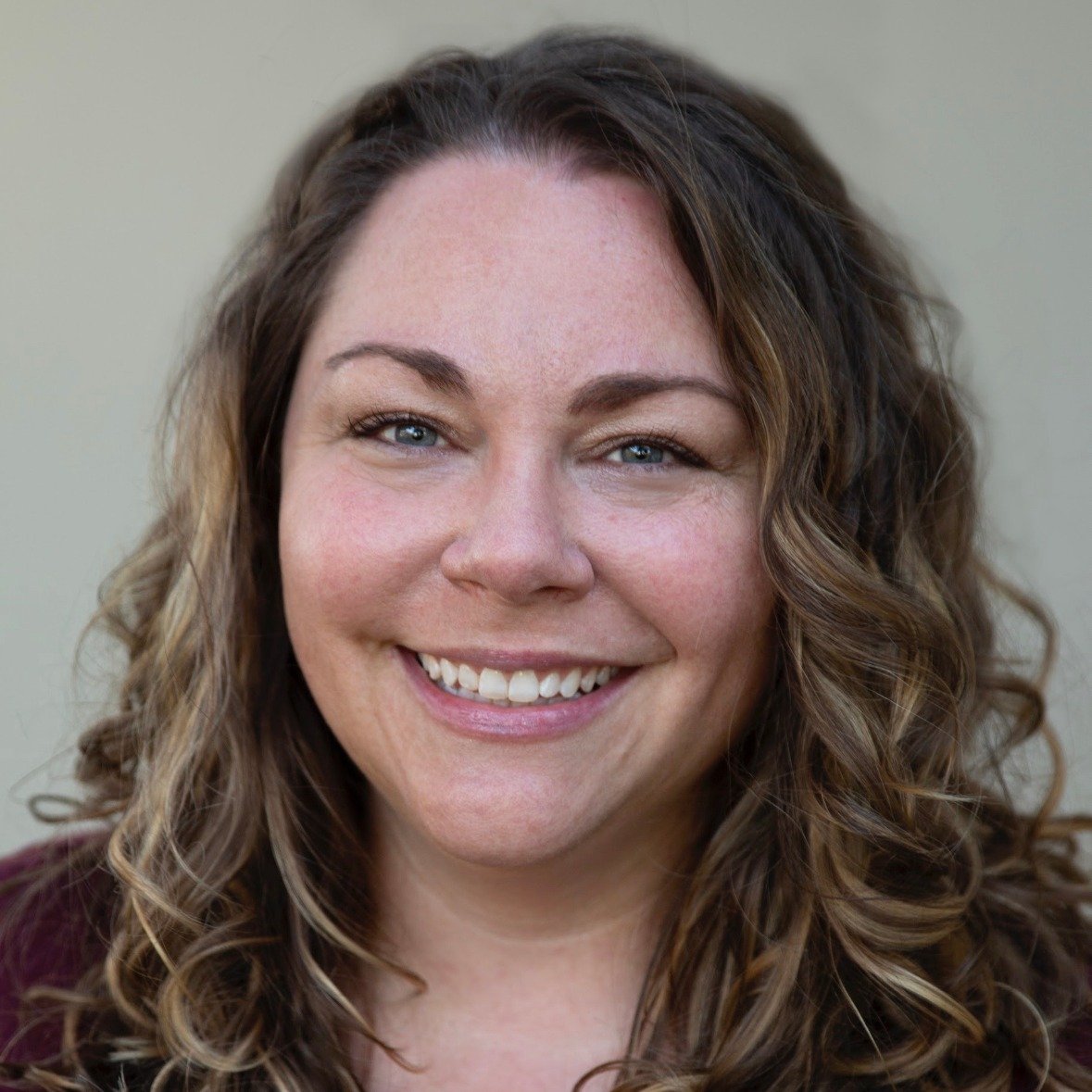 Close-up of a smiling woman with wavy brown hair and blue eyes, wearing a dark top, against a plain light background.