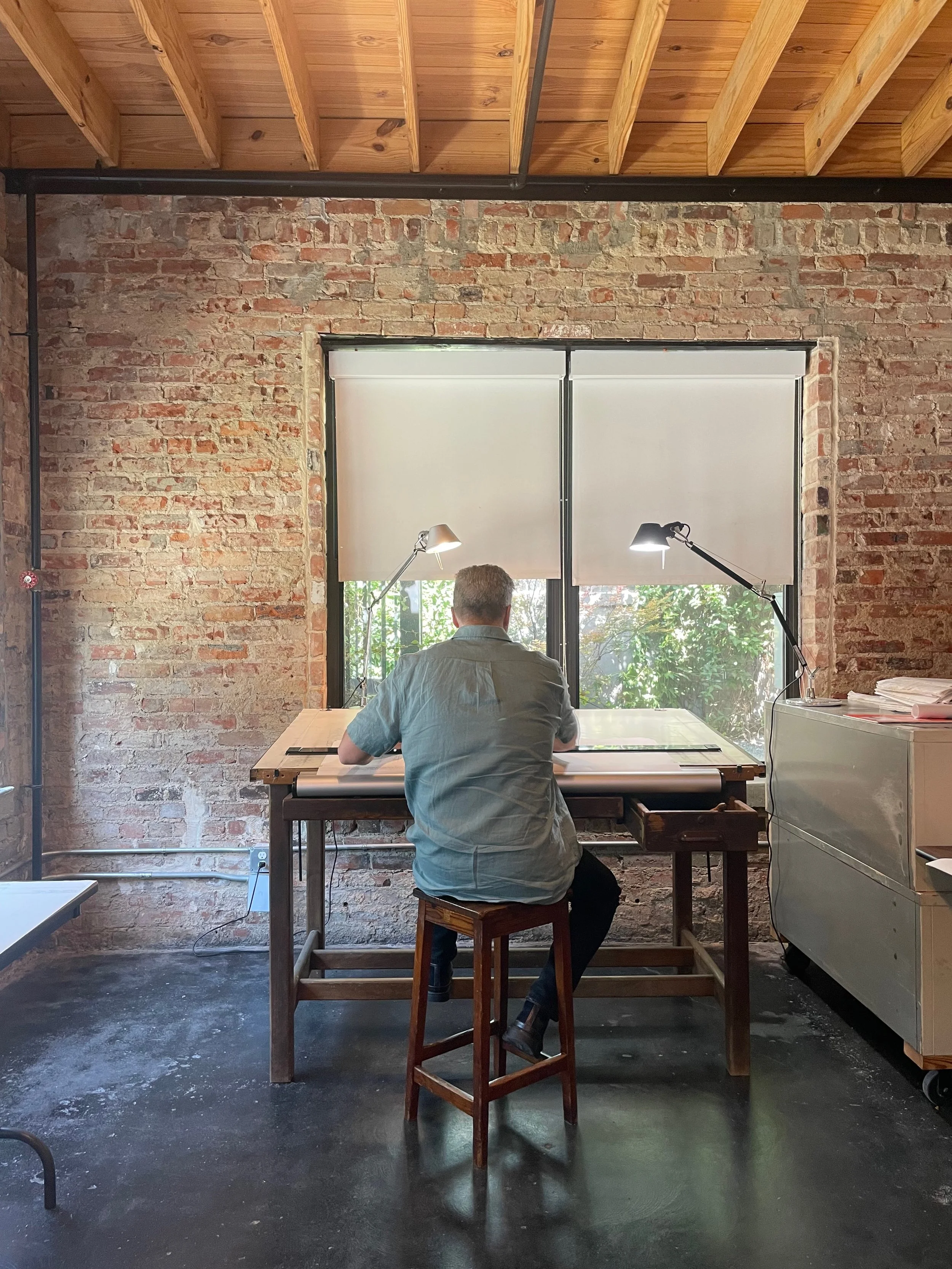 A man with gray hair, wearing a short-sleeved button-up shirt, sitting at a drafting table in front of a large window with white blinds, brick wall background, two adjustable desk lamps, and a white cabinet with papers on top.