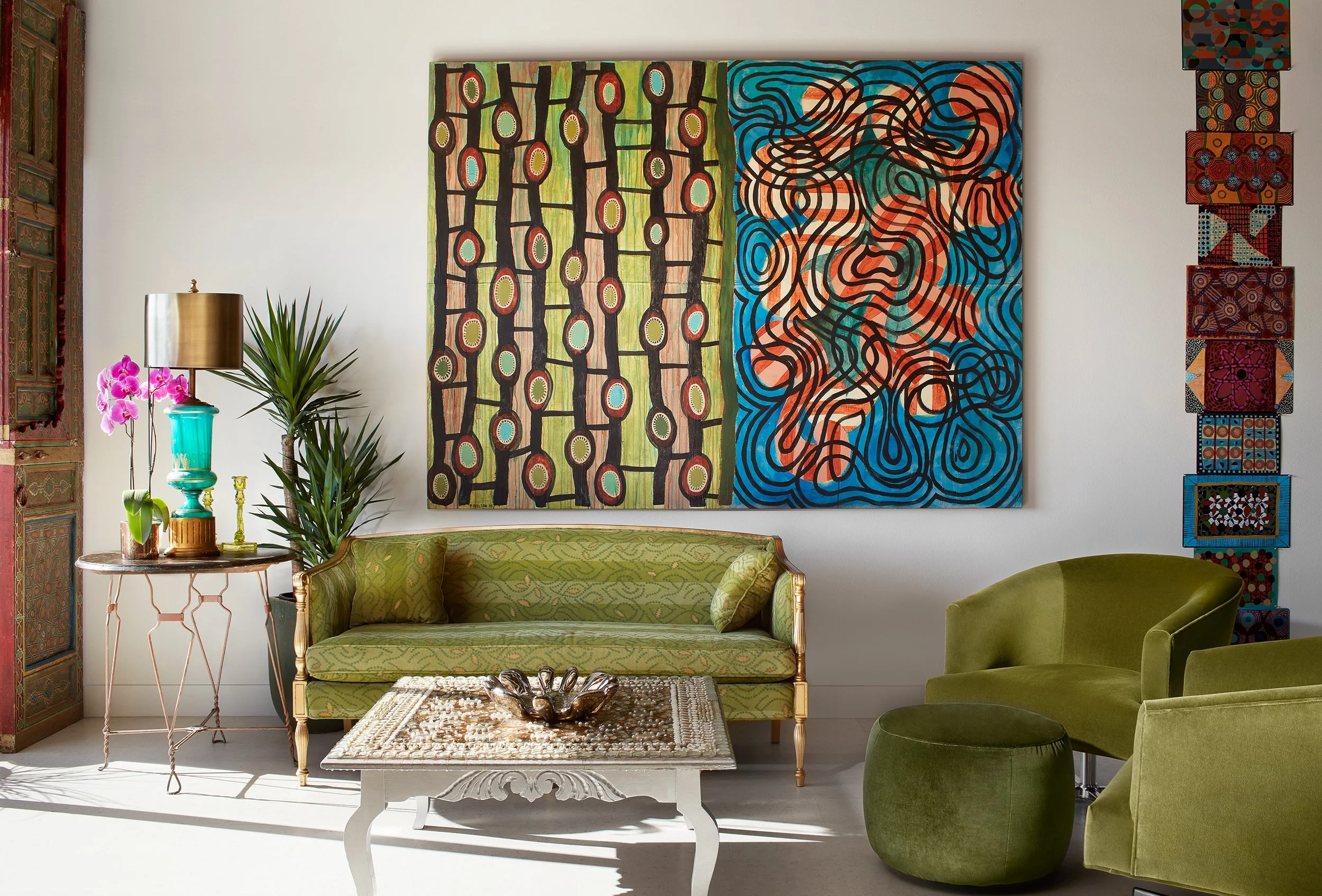 Interior of a living room featuring abstract paintings on a white wall, a green vintage sofa, a round coffee table with a decorative sculpture, and a side table with a gold lamp, a teal vase, and pink orchids. There is a green armchair and a matching ottoman nearby, and a decorative wooden door on the left.