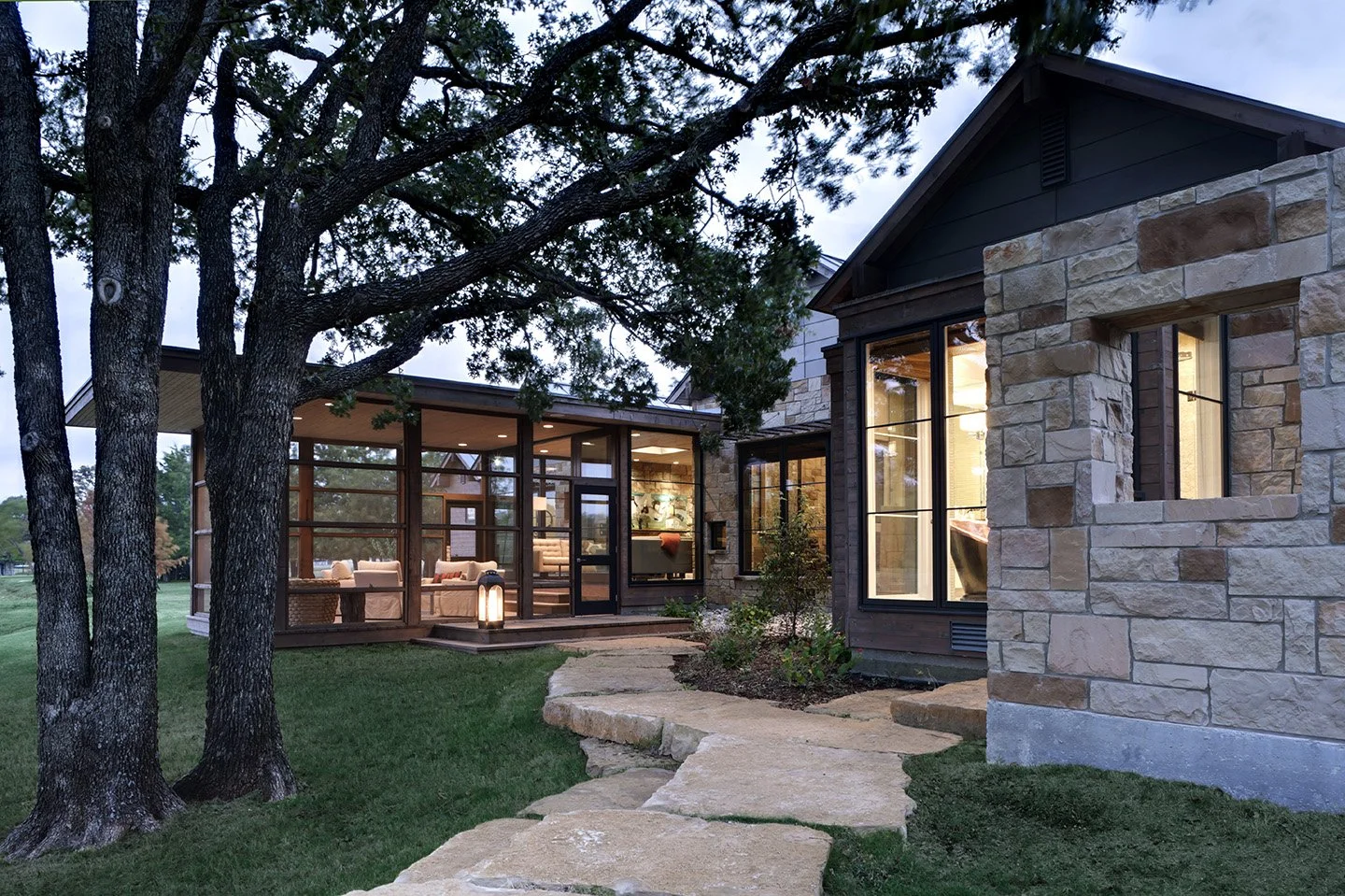 Exterior view of a modern house with a glass sunroom, stone and wood siding, illuminated from inside, with a stone pathway leading to the entrance, surrounded by trees and a green lawn during twilight.