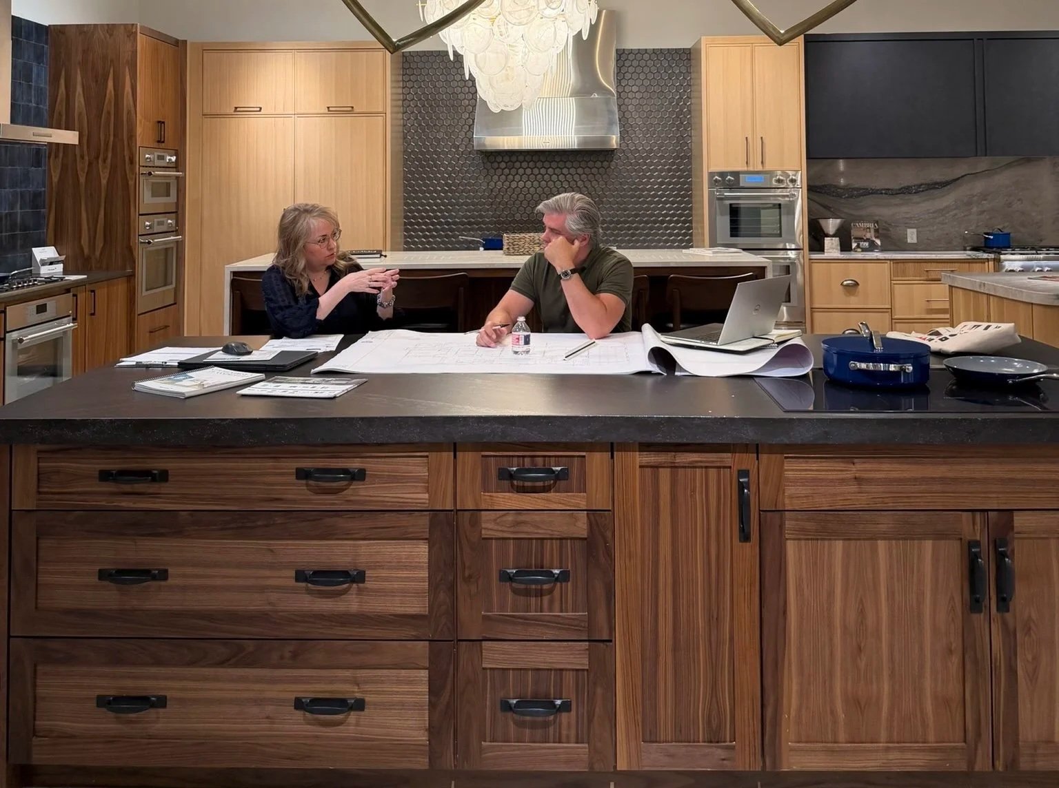 Two people sitting at a large kitchen island table discussing, with documents, blueprints, a laptop and a water bottle on the table, in a modern kitchen with wooden cabinets and stainless steel appliances.