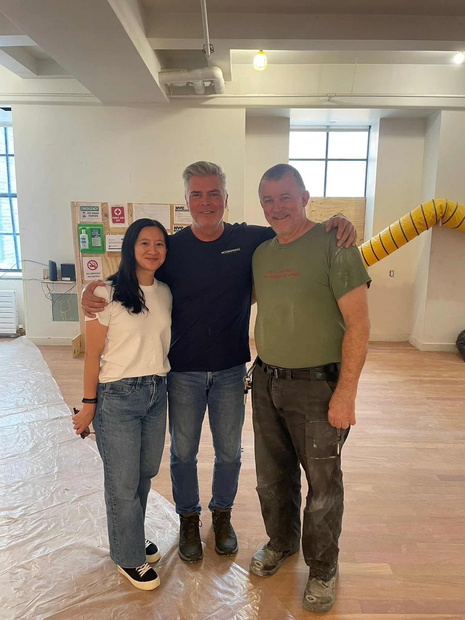 Three people standing together indoors, smiling, with a woman on the left and two men in the middle and right. They appear to be at a construction site or renovation area, with a plastic sheet on the floor and signs on a notice board in the background.