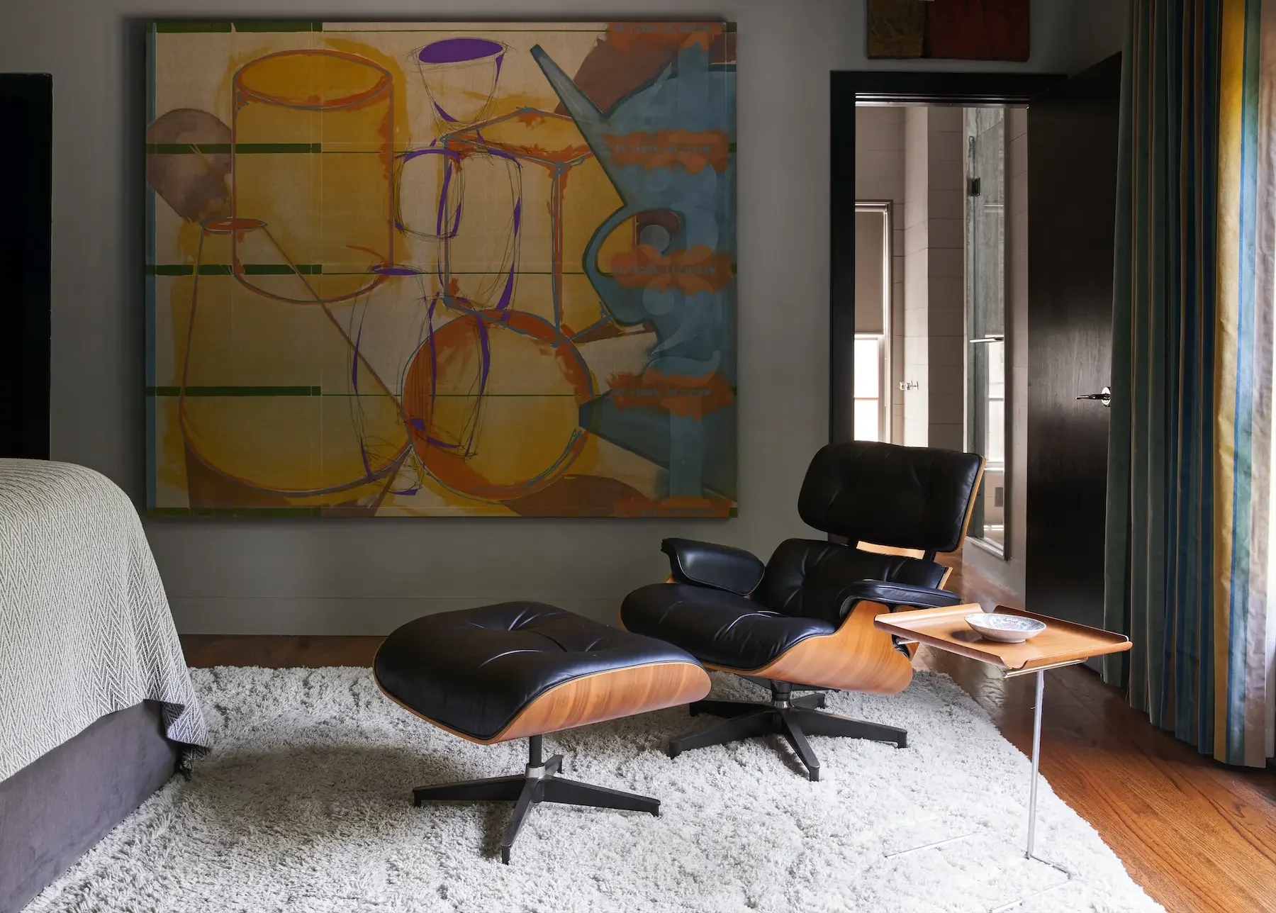 Living room with modern furniture, including a black leather lounge chair with matching ottoman, a wooden side table, and a patterned armchair. Large abstract artwork hangs on the wall, and there is a white shag rug on the hardwood floor. A doorway leads to a kitchen with tile walls, a window, and a concrete countertop. Blue and yellow striped curtains are partially visible on the right.