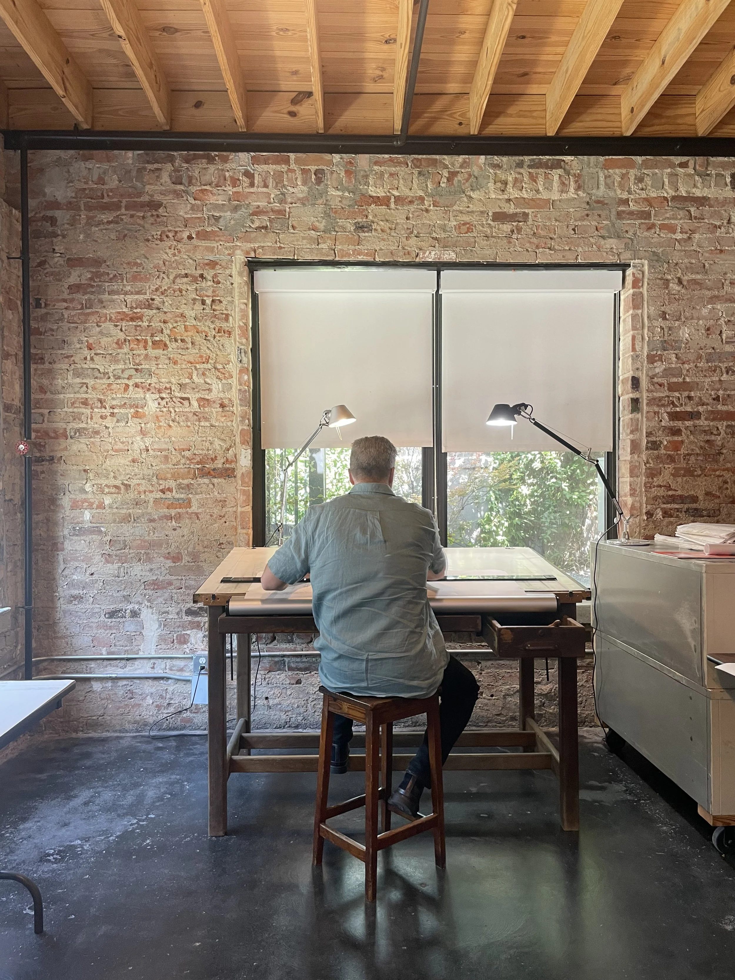 A man with short gray hair sitting at a drafting table in an industrial-style room with brick walls and a wooden ceiling. The window behind him has white roller shades, and two adjustable desk lamps are positioned on the table, illuminating his workspace.