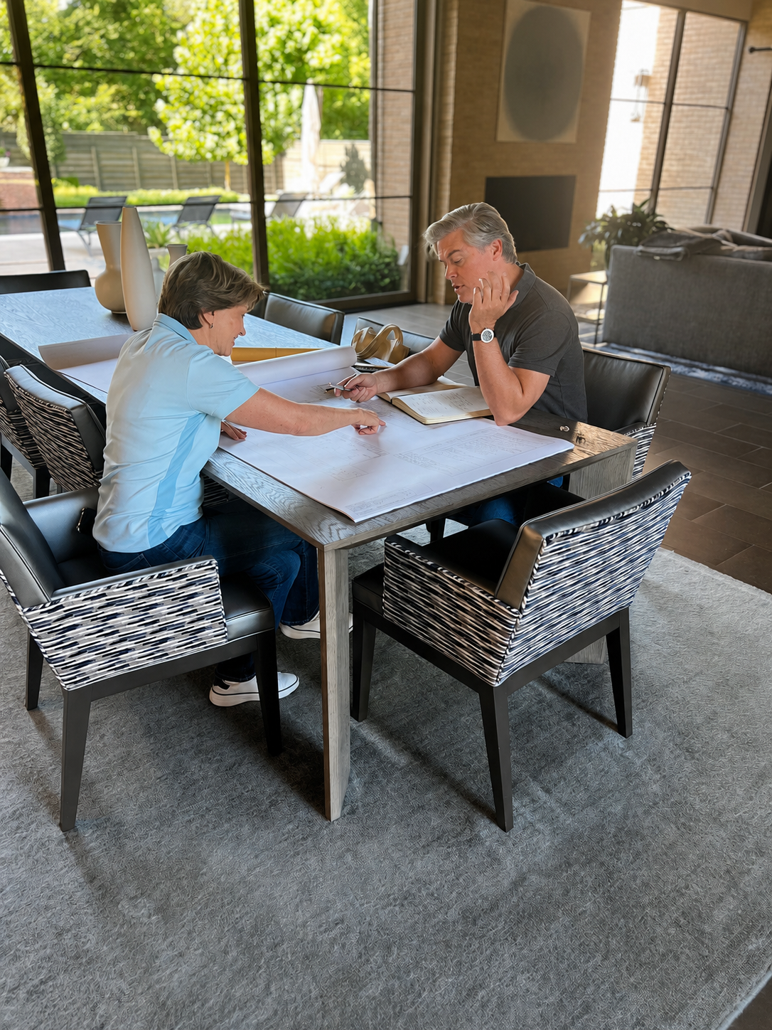 A young boy and an older man are sitting at a dining table with large architectural plans spread out between them. The boy is pointing at the plans, and the man is looking at them while resting his chin on his hand, with an open notebook in front of him. The room has large windows revealing an outside patio with greenery.