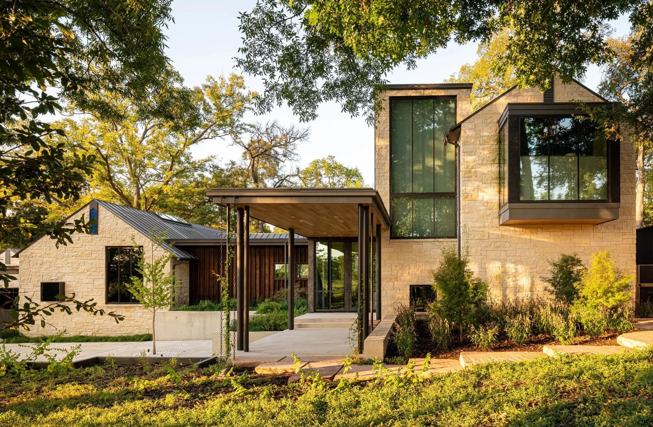 A modern house with large glass windows and a stone exterior, surrounded by trees and a landscaped yard.
