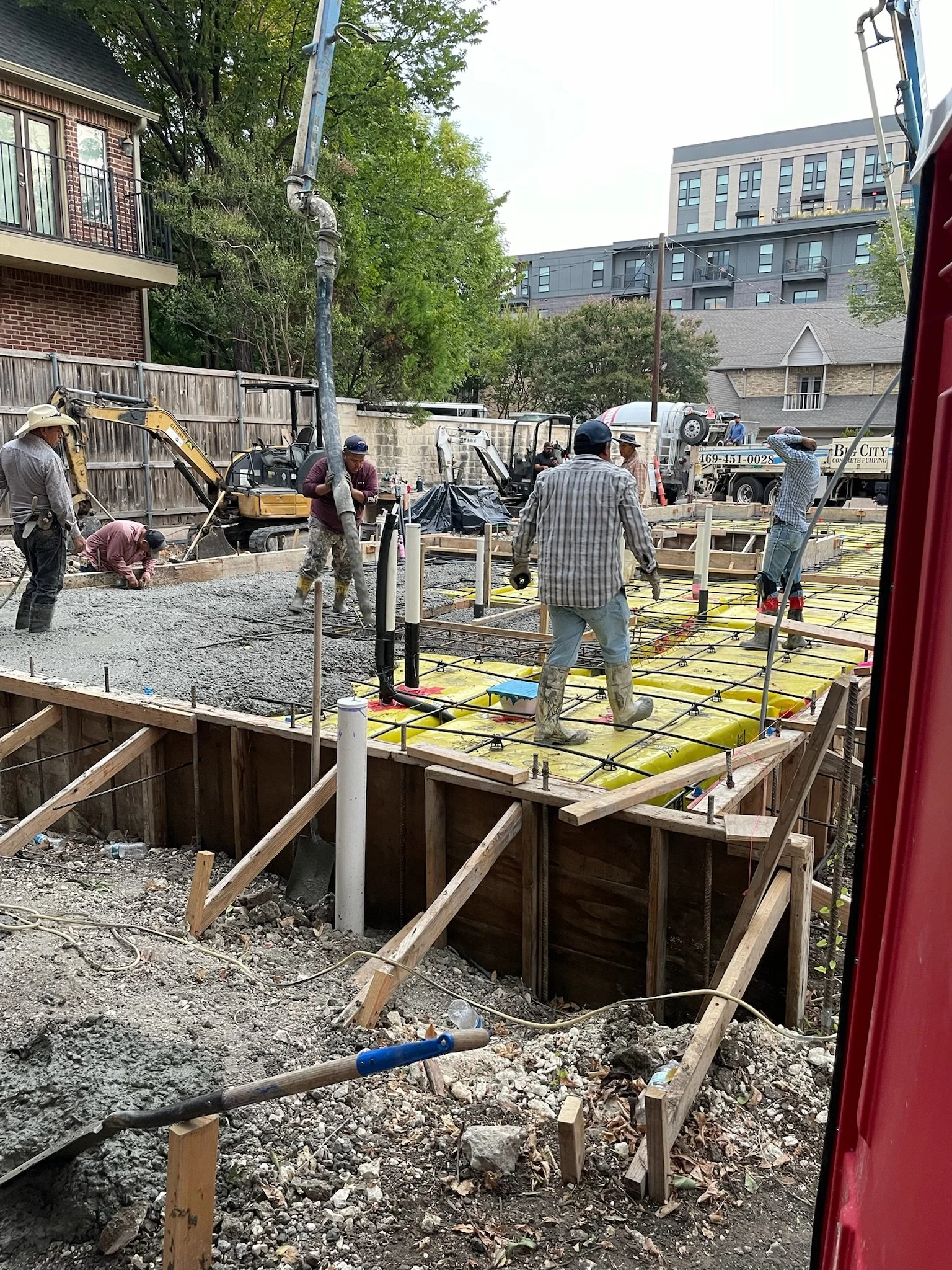 Construction workers pouring concrete on a building site with wooden formwork and rebar, heavy machinery, and a water truck in the background.