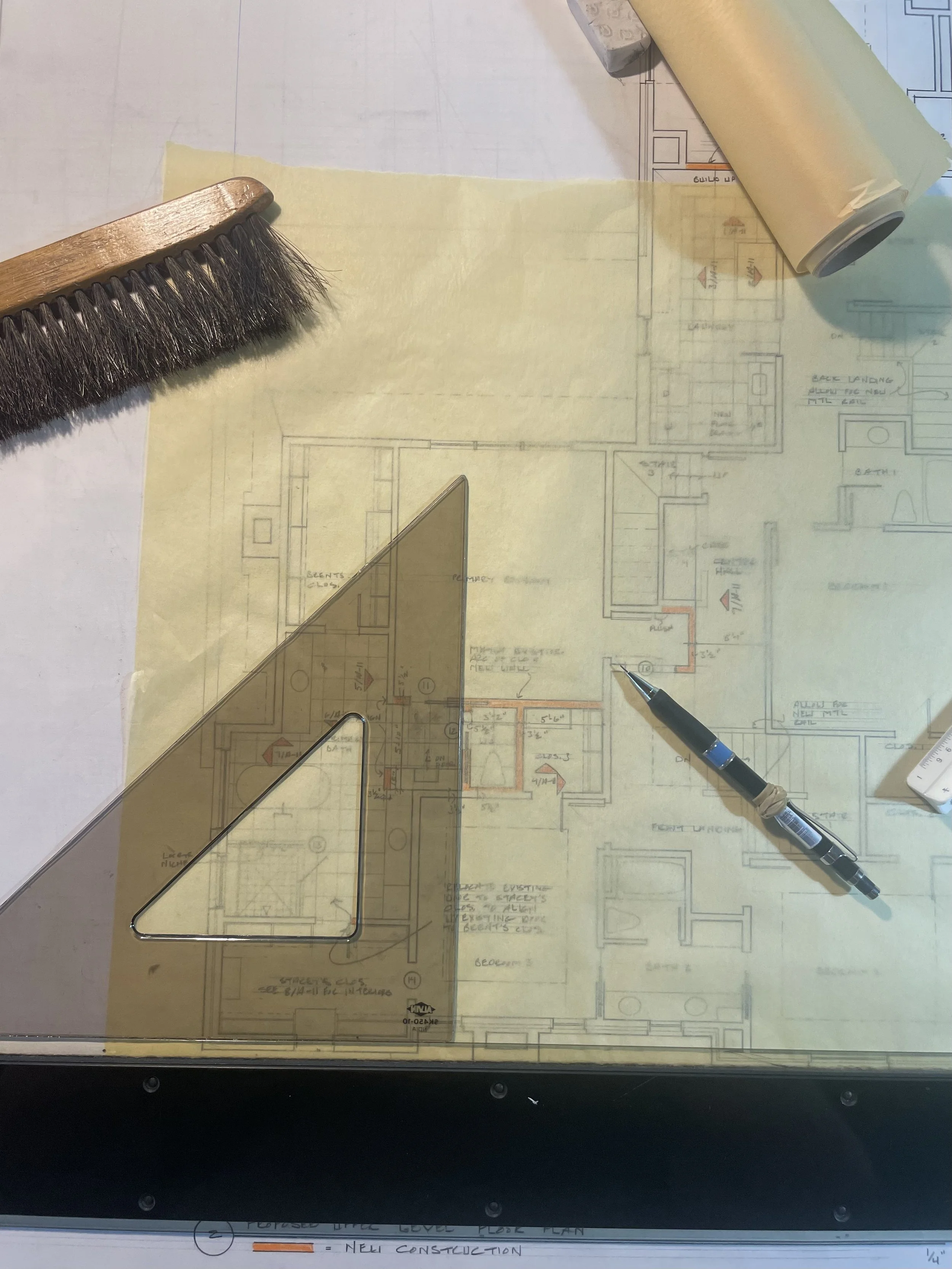 Architectural blueprint of a building layout with tools including a paintbrush, a pencil, a metal triangle ruler, and a rolled-up paper on top.