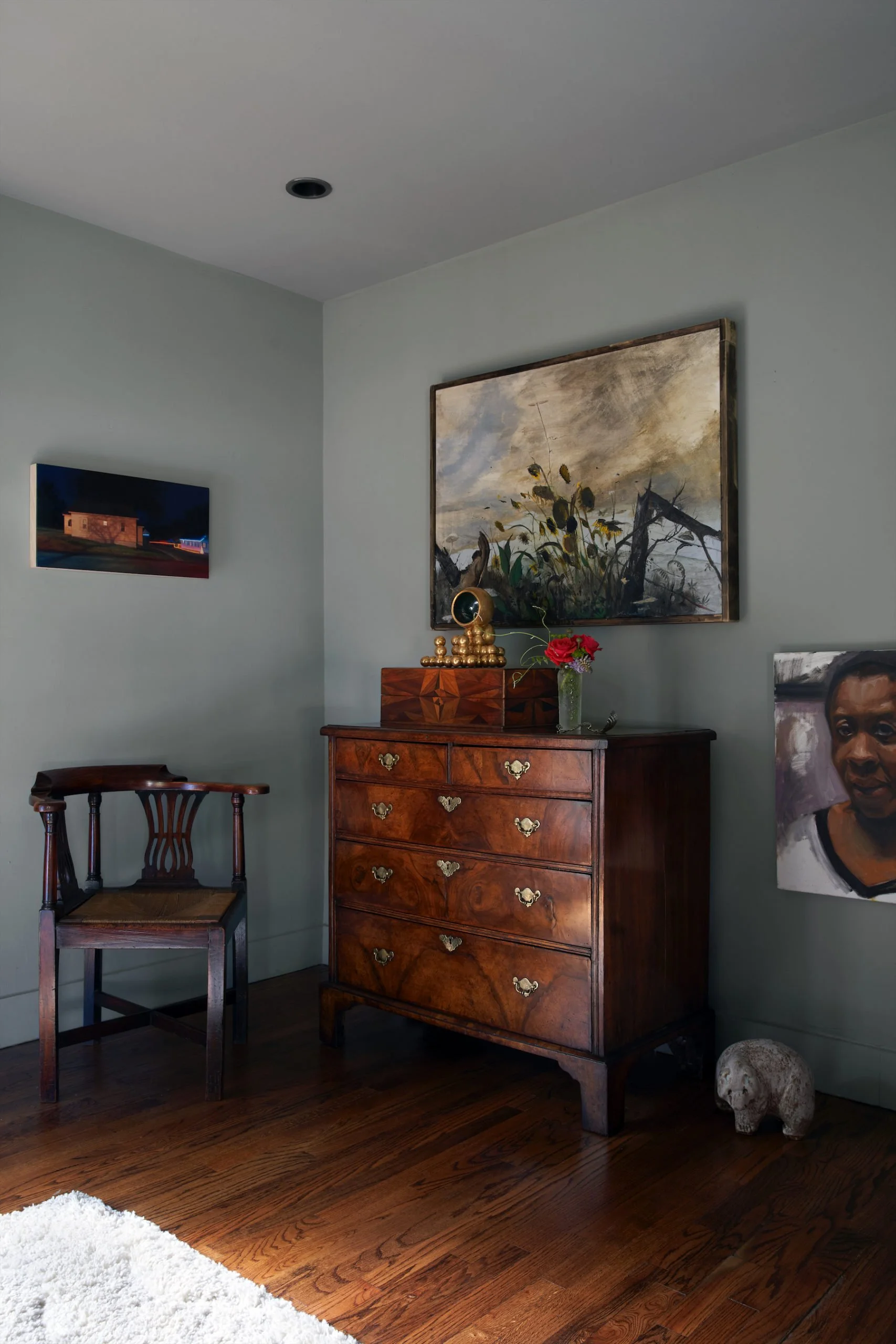 A corner of a room with wooden flooring, a wooden chair, a vintage wooden dresser with brass handles, a small woman skull, a glass vase with red flowers, two framed paintings on the walls, a decorative golden object on the dresser, and a piece of white fluffy rug on the floor.