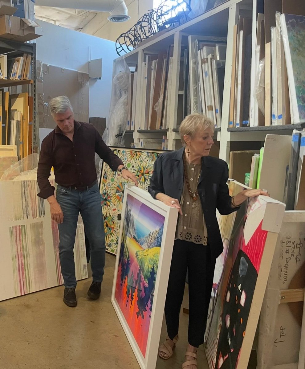 Two people look at paintings in an art store or gallery. The woman is holding a colorful landscape painting, and the man is looking at paintings on the wall. Shelves filled with framed artwork are in the background.