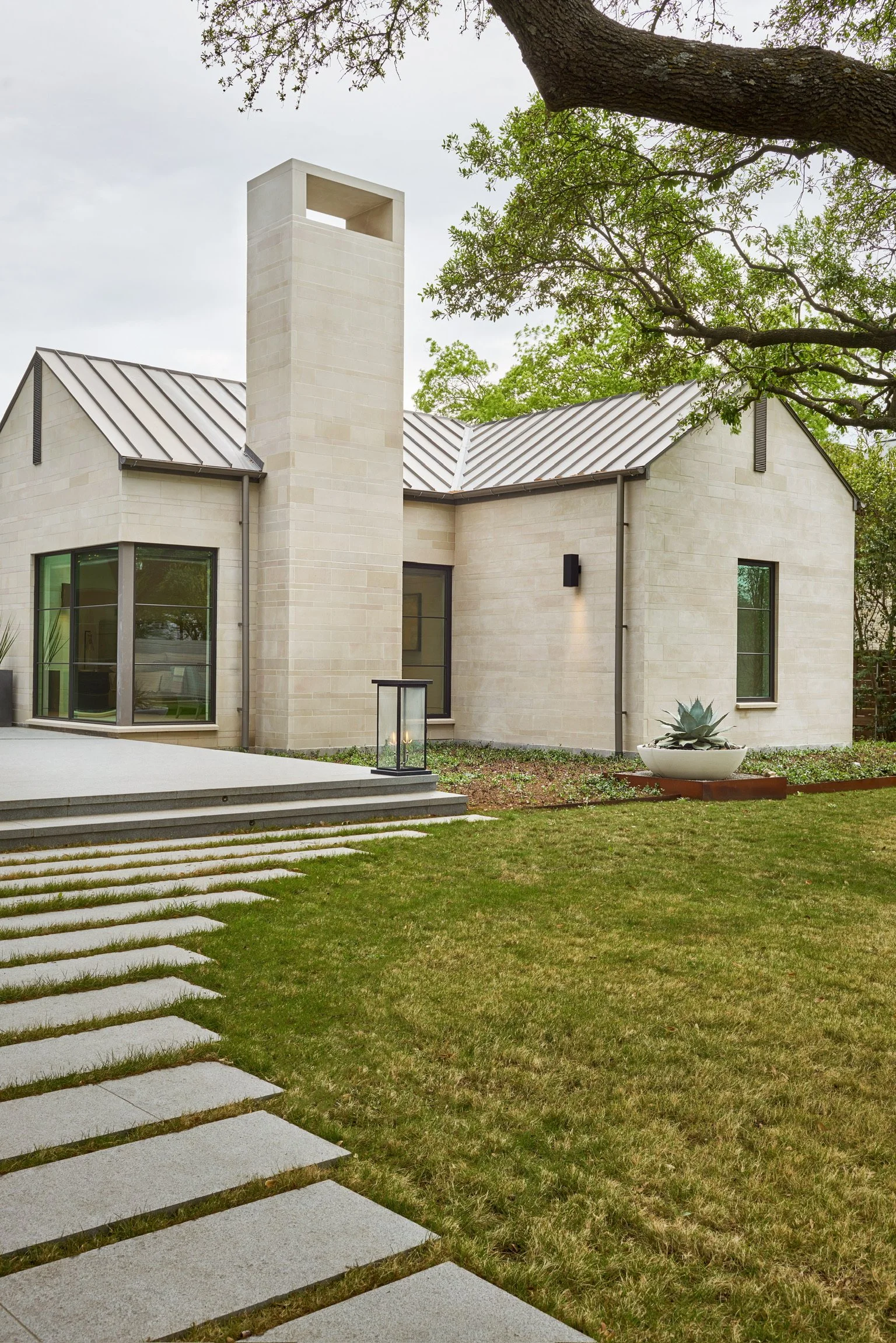 A modern house with a light-colored brick exterior, large windows, a metal roof, and a chimney. The house is surrounded by greenery, including a tree with green leaves, a grassy lawn, and a pathway made of stepping stones leading to the house. There is a lantern near the pathway and a large white planter with a succulent plant.