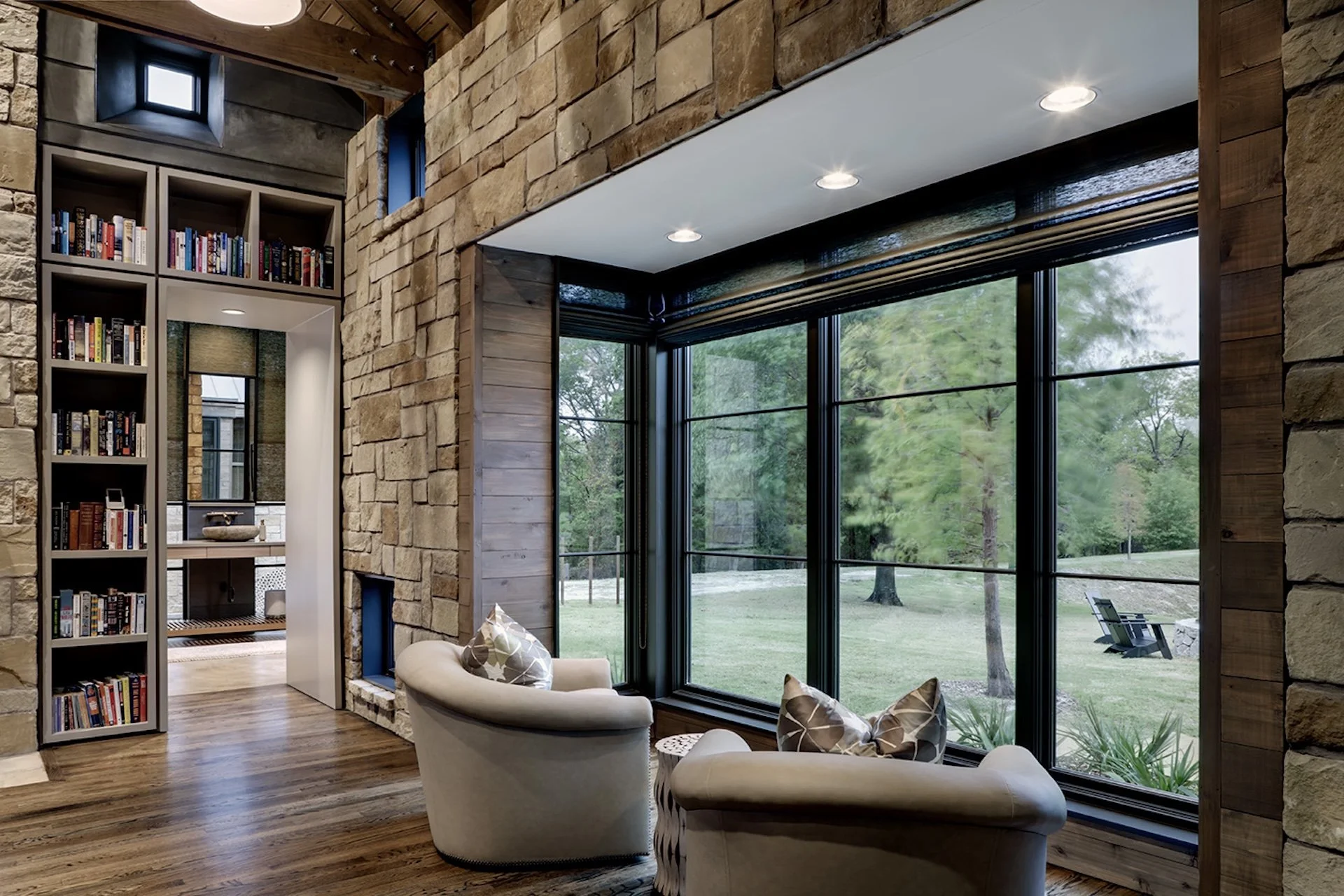 Living room with large windows overlooking a green outdoor area, stone and wood wall accents, a cozy seating area with a white armchair and patterned pillows, and a bookshelf visible through a doorway.