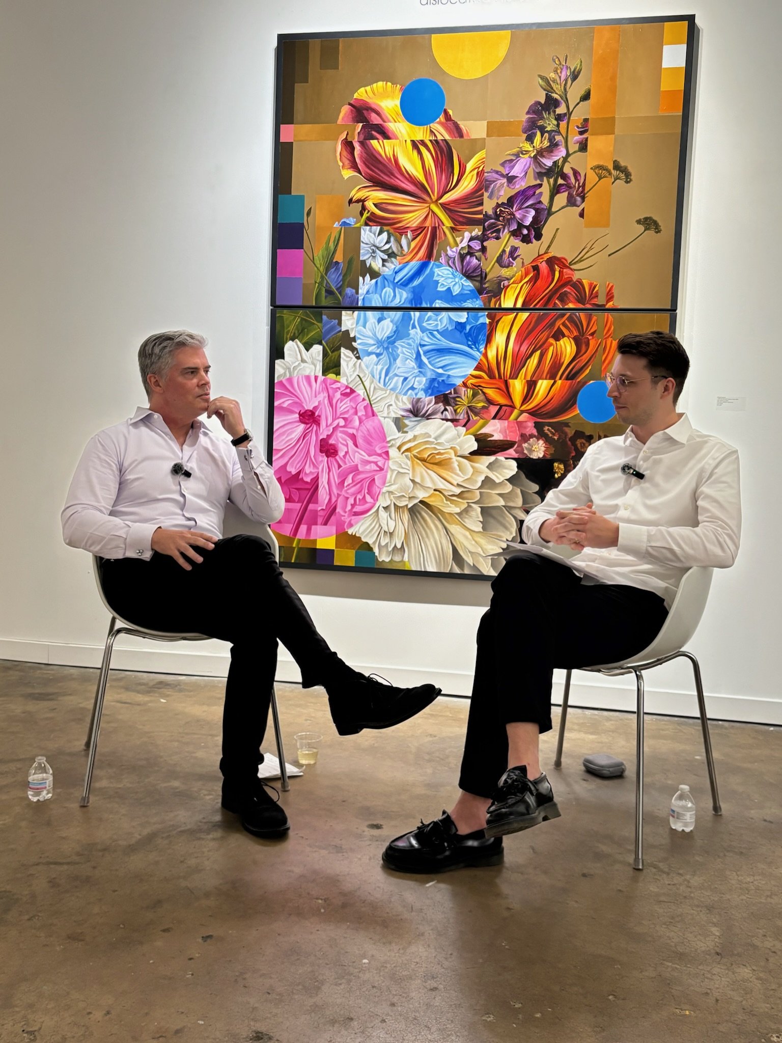 Two men dressed in white shirts are seated in chairs having a conversation in an art gallery. The background features a colorful, abstract floral painting.