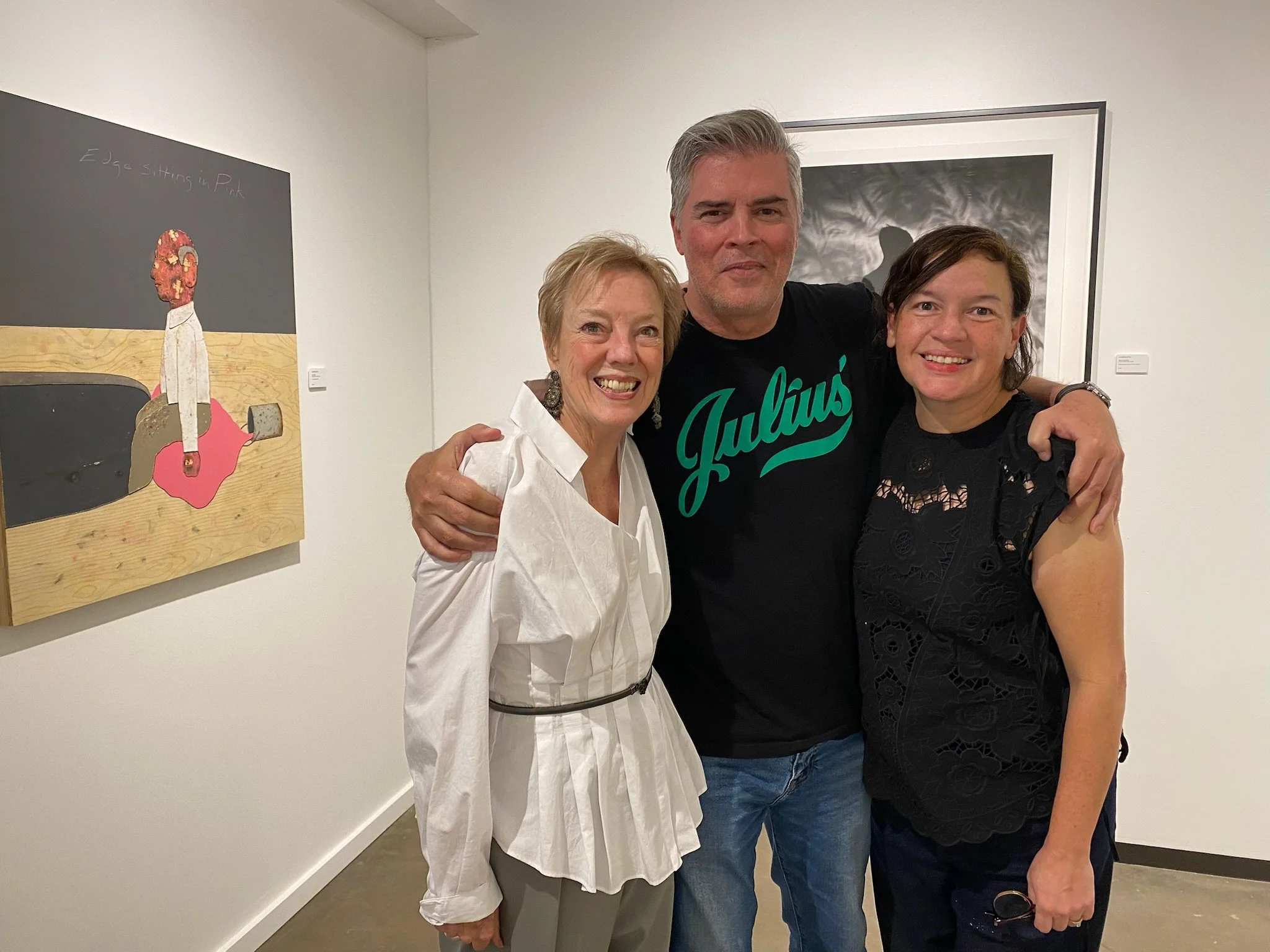 Three people, two women and a man, smiling and standing close together in an art gallery, with artwork on the wall behind them.