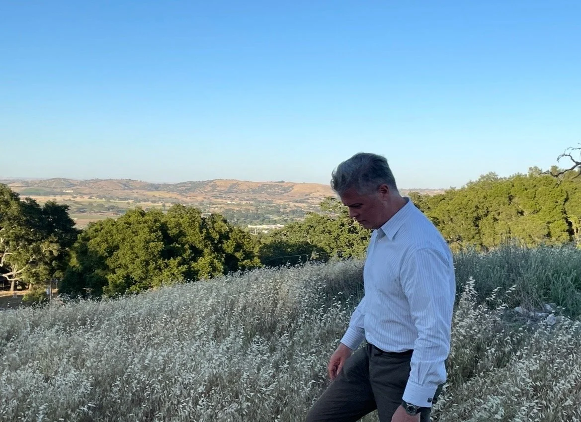 A man in a white shirt standing in a field of tall grass with trees and hills in the background under a clear blue sky.