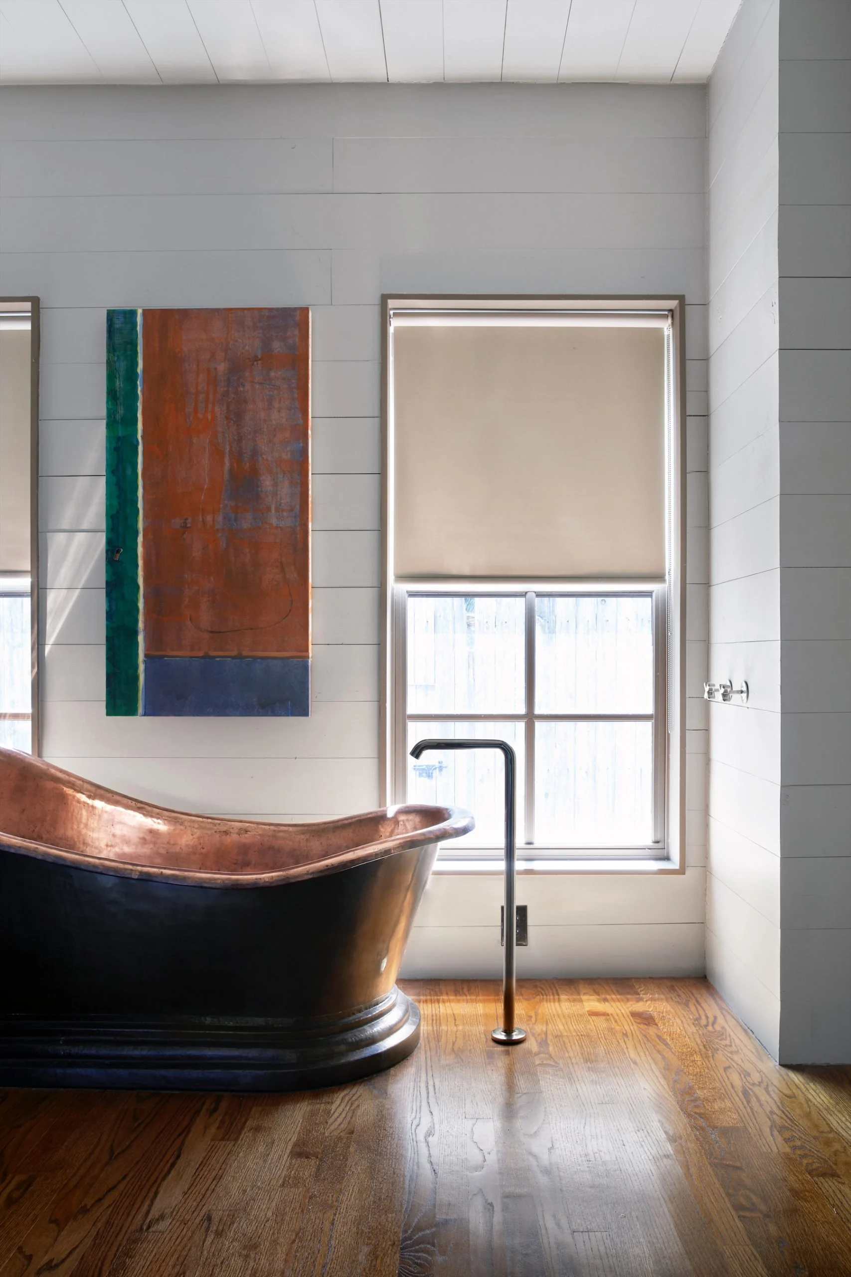 A bathroom with a copper bathtub, white shiplap walls, a window with a beige roller blind, and wooden flooring.