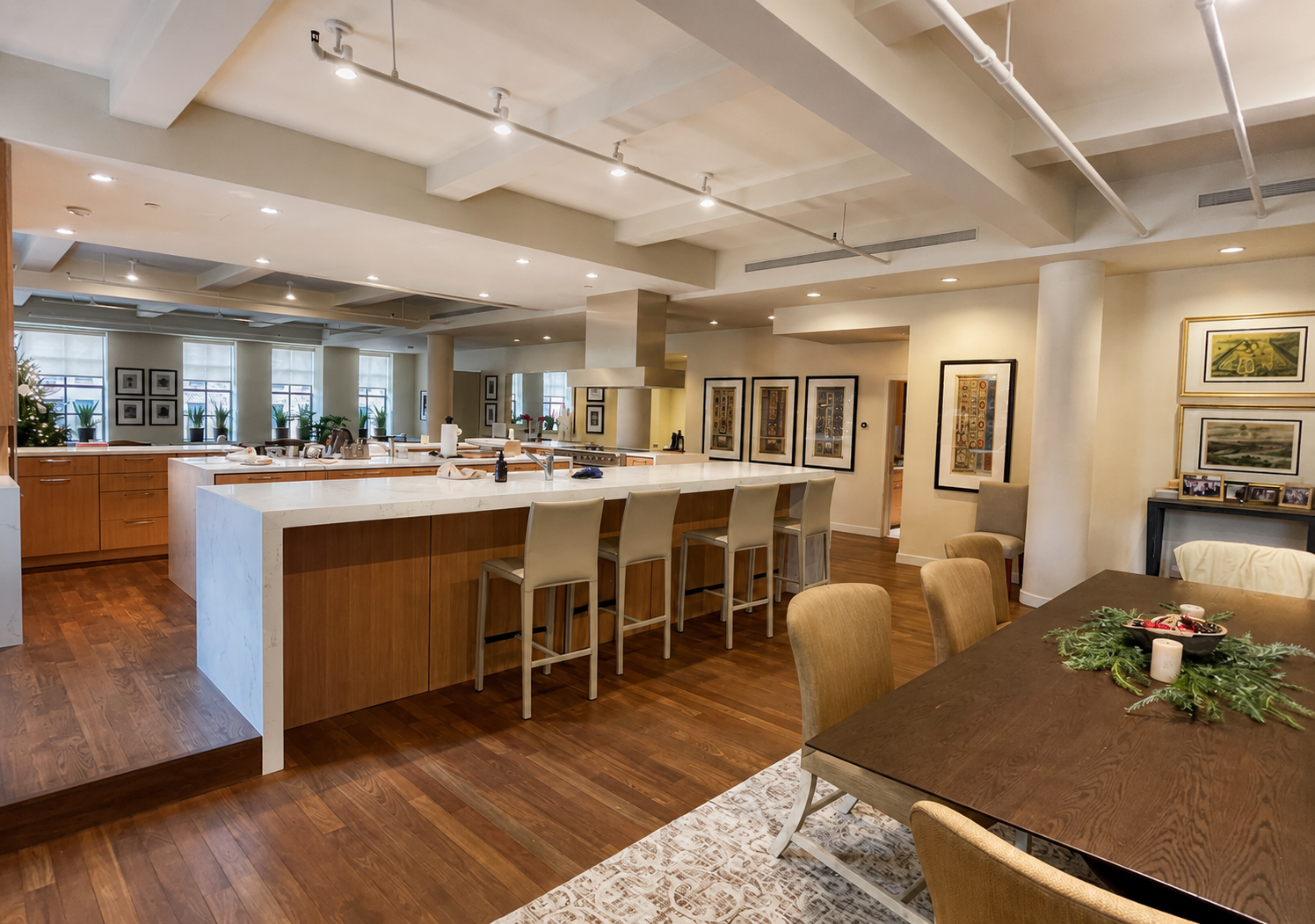 A spacious open-concept kitchen and dining area with wooden floors, a large kitchen island with a white countertop, beige bar stools, a cooking range with a hood, and decorated walls with framed artwork. There are windows with potted plants and Christmas decorations, and a dining table with holiday decorations.