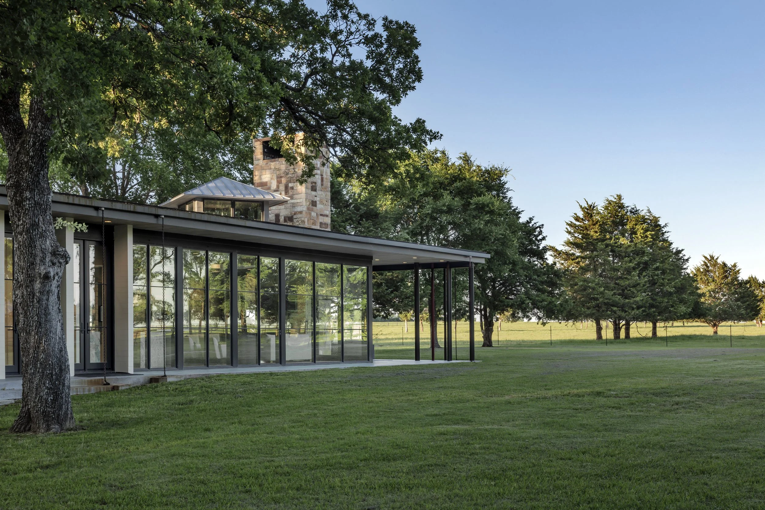 Modern house with large glass windows and a metal roof, surrounded by green grass and tall trees, under a clear blue sky.