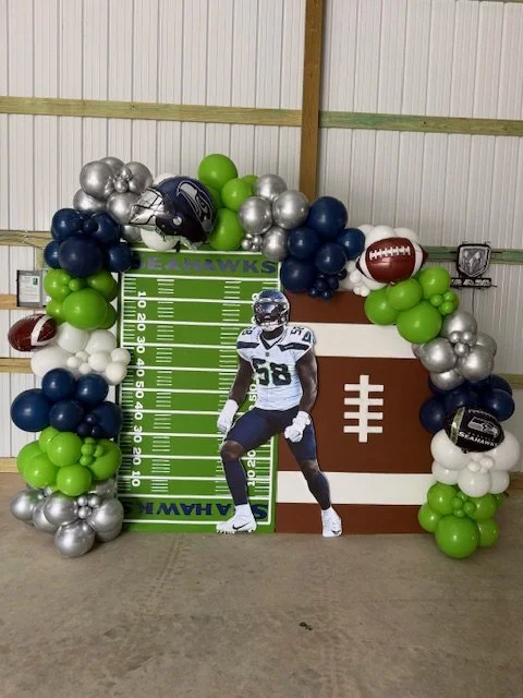 Football-themed party backdrop with a balloon arch, a cardboard cutout of a football player, and a chalkboard with a football field design.