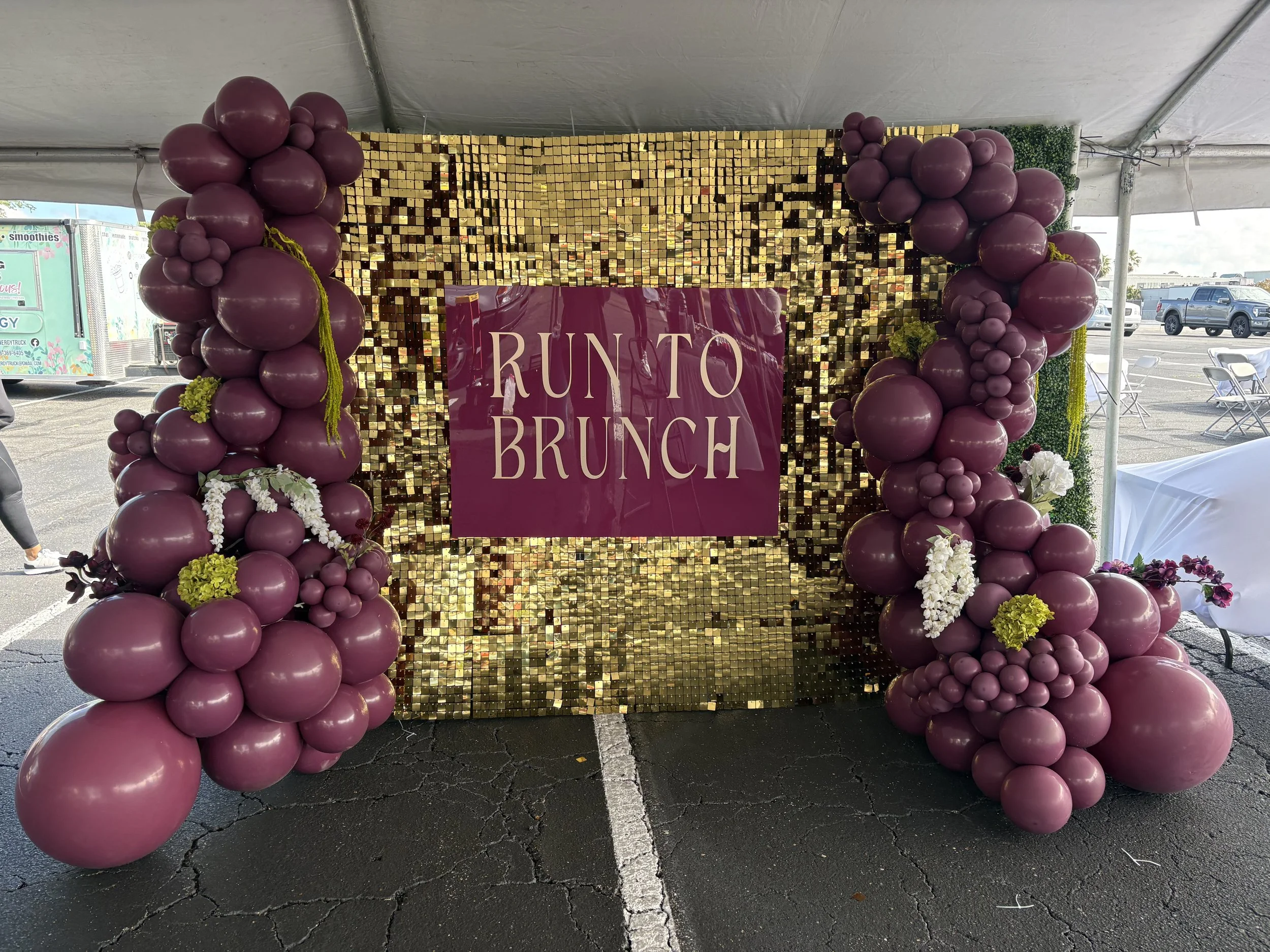 A decorative backdrop for an event with purple balloon clusters surrounding a golden sequin wall and a magenta banner that reads "Runto Brunch."