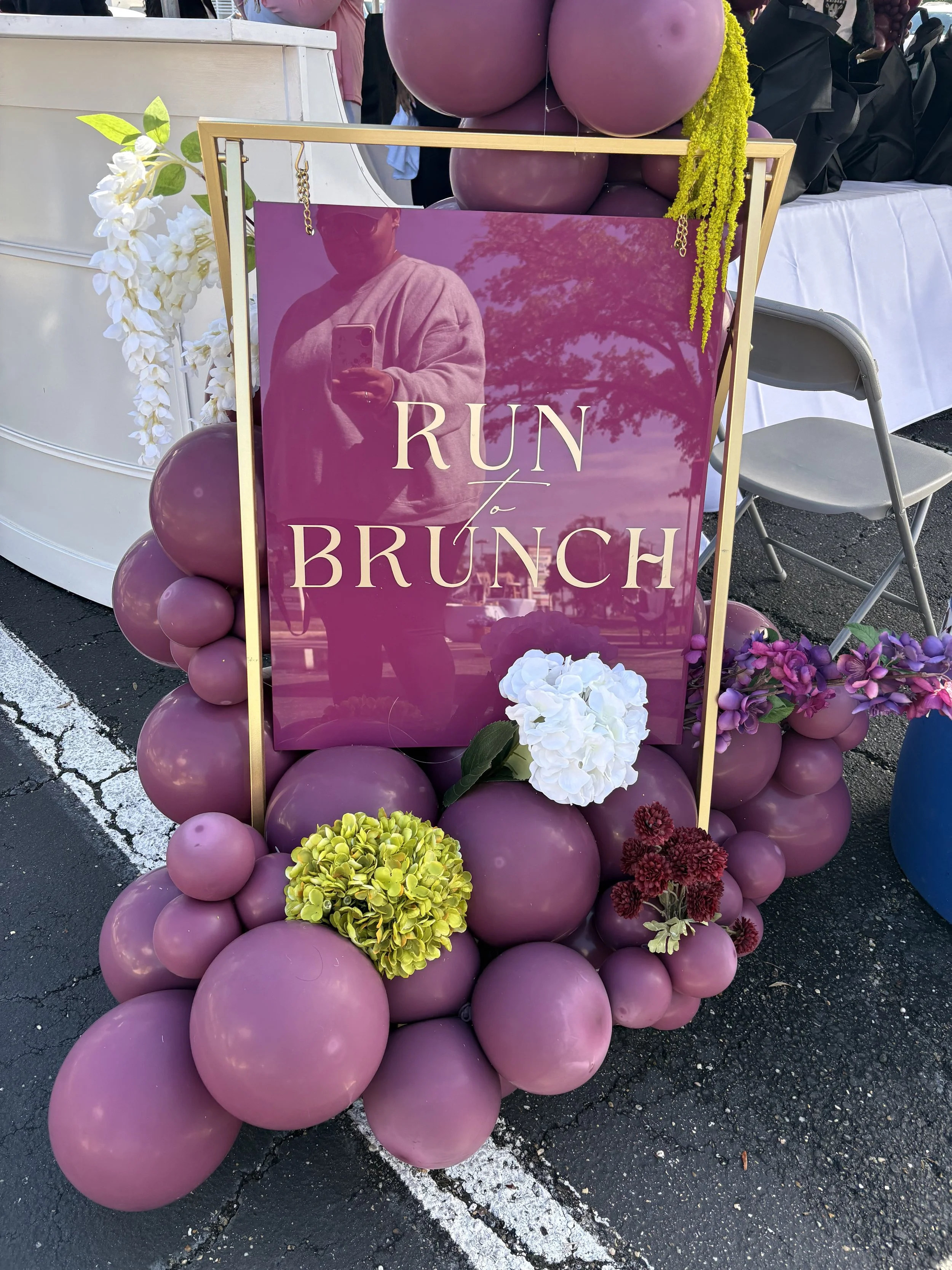A decorative board with the words 'Run to Brunch' written on a mirrored surface, surrounded by pink and purple balloons and flower arrangements.
