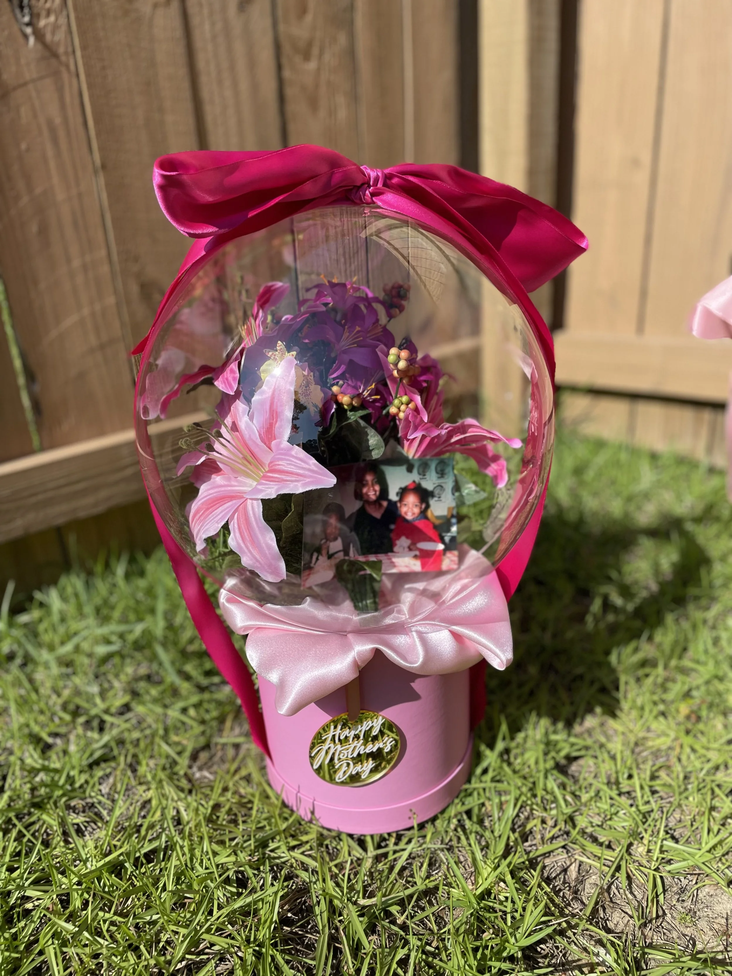 A Mother's Day flower bouquet with pink lilies and purple flowers in a pink wrapping, placed on grass in front of a wooden fence.