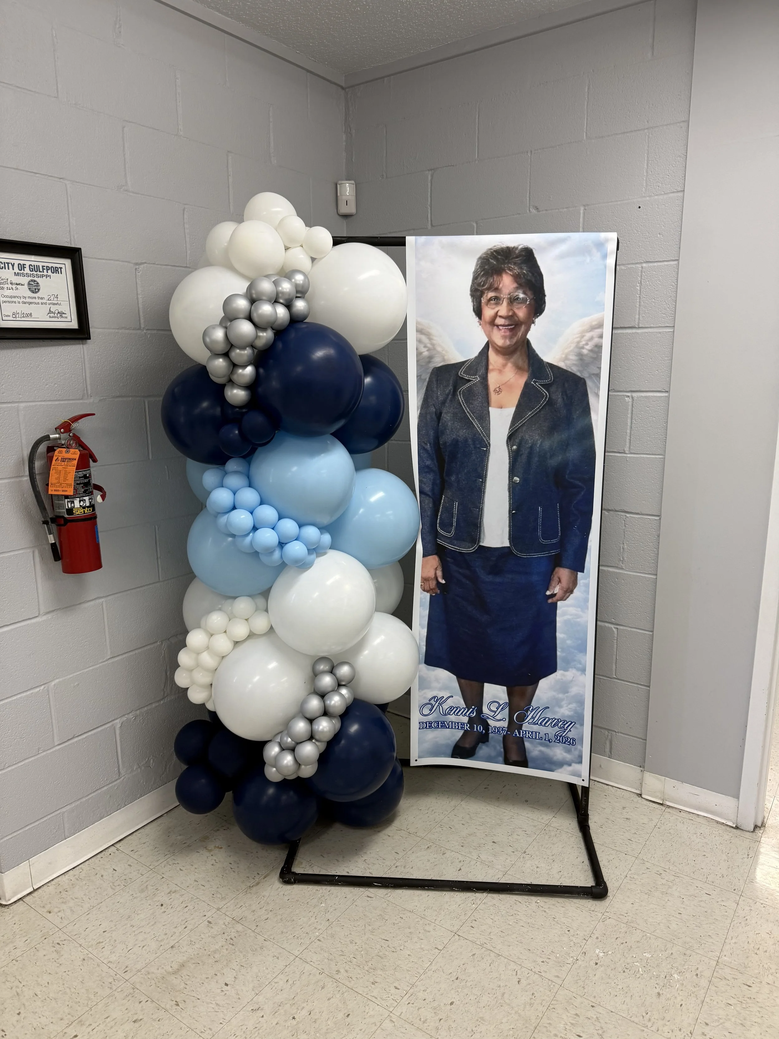 Funeral display with a balloon arrangement in shades of white, gray, light blue, and dark blue next to a poster featuring a woman with angel wings, dressed in a dark blazer and white top, with text indicating her name and dates of birth and death.