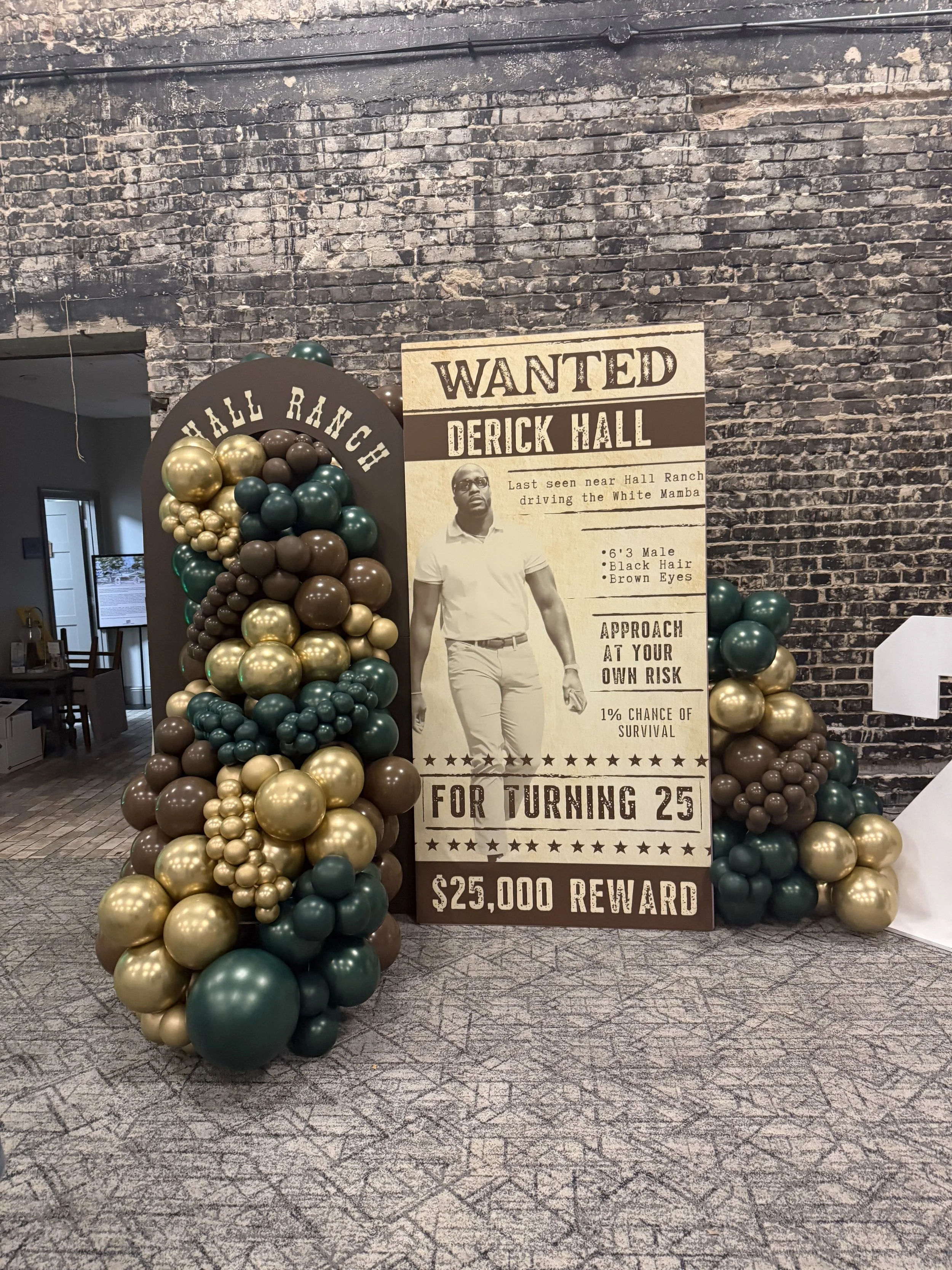 A wanted poster for Derick Hall, featuring a photo of a man in sunglasses and a white polo shirt, with a background of dark brick wall and gold and green balloons arranged around the poster. The poster states Derick Hall is wanted for driving the White Mamba near Hall Ranch, has a height of 6'3", black hair, and brown eyes, with a $25,000 reward.