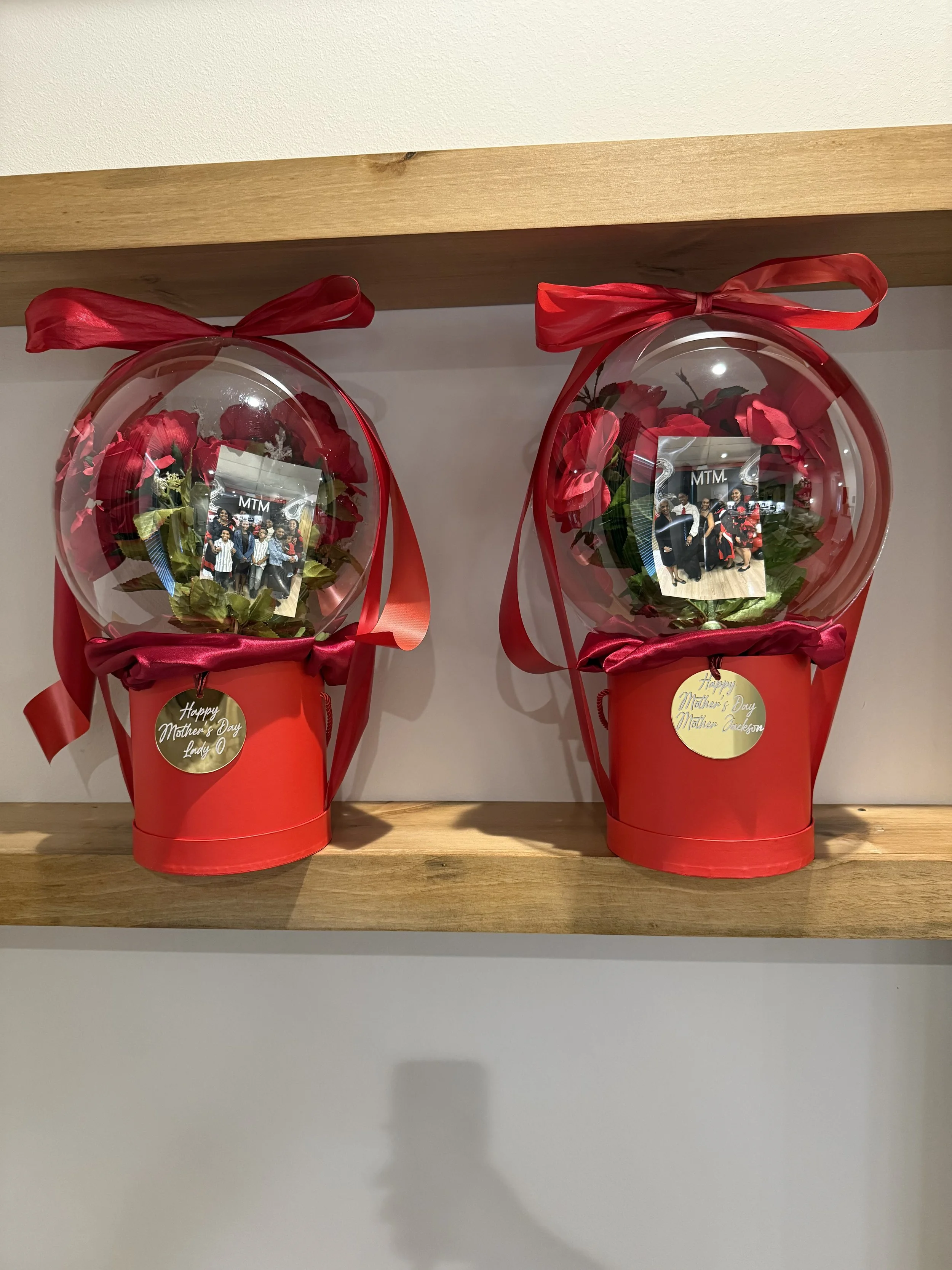 Two decorative flower arrangements in red buckets with transparent domes, red ribbons on top, flowers and a photo inside each, placed on a wooden shelf.