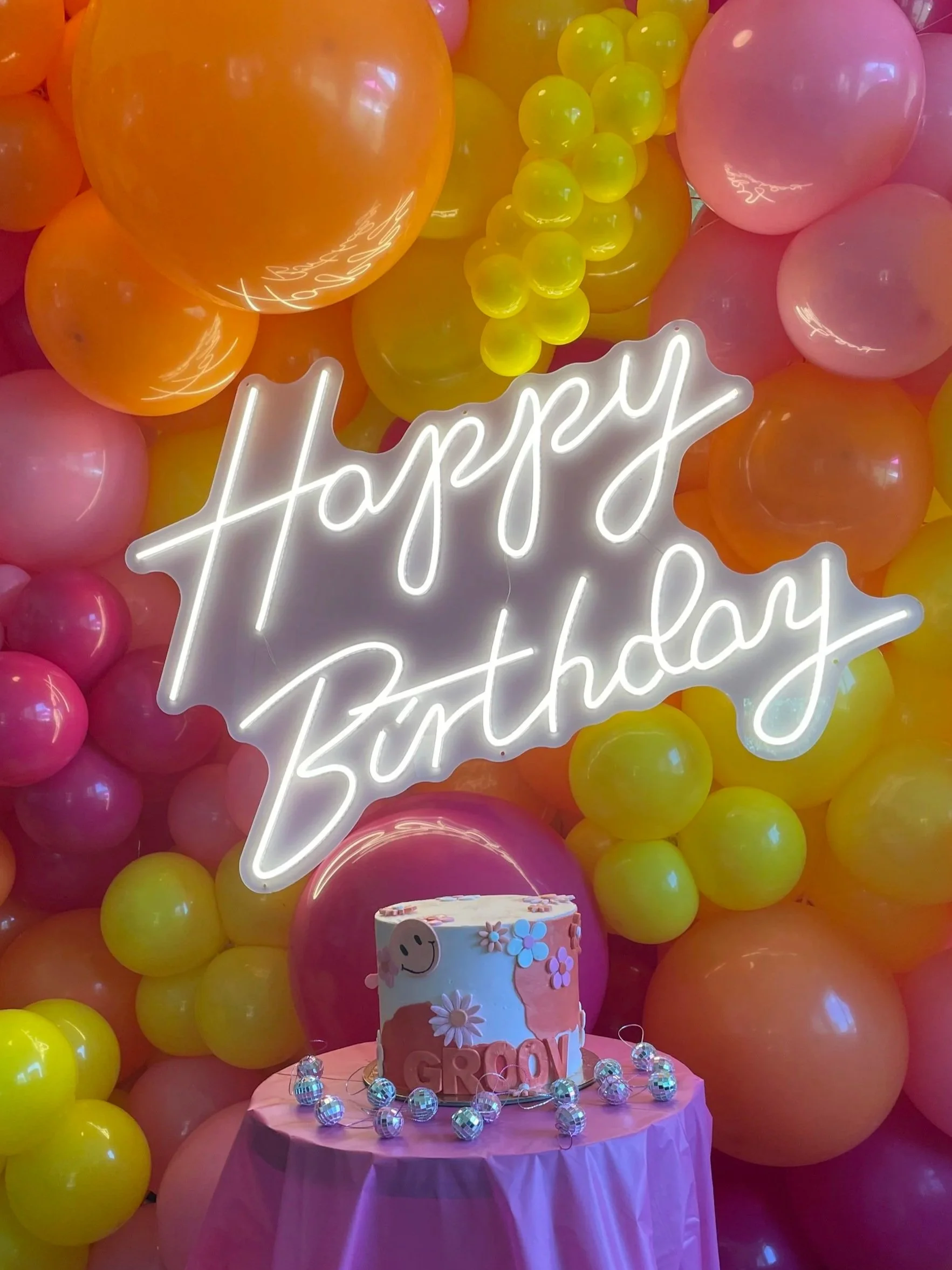 Colorful birthday party decoration with pink, yellow, and orange balloons in the background, a neon sign that reads 'Happy Birthday,' a cake with a smiley face, daisies, and the word 'GIRL,' and small reflective decorative balls surrounding the cake on the table.