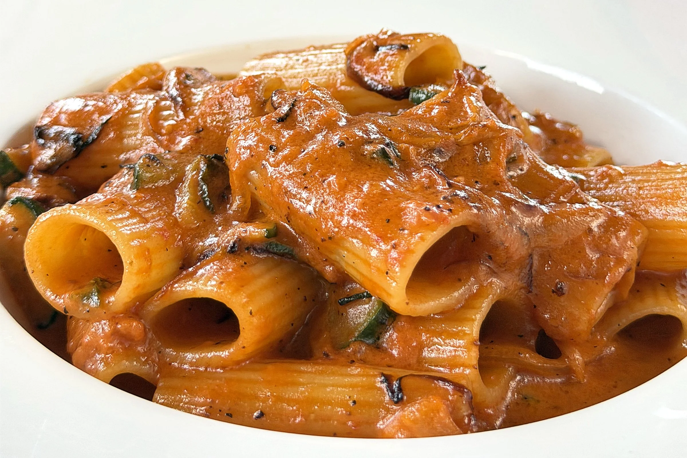 Close-up of cooked rigatoni pasta with tomato sauce, mushrooms, and herbs on a white plate.