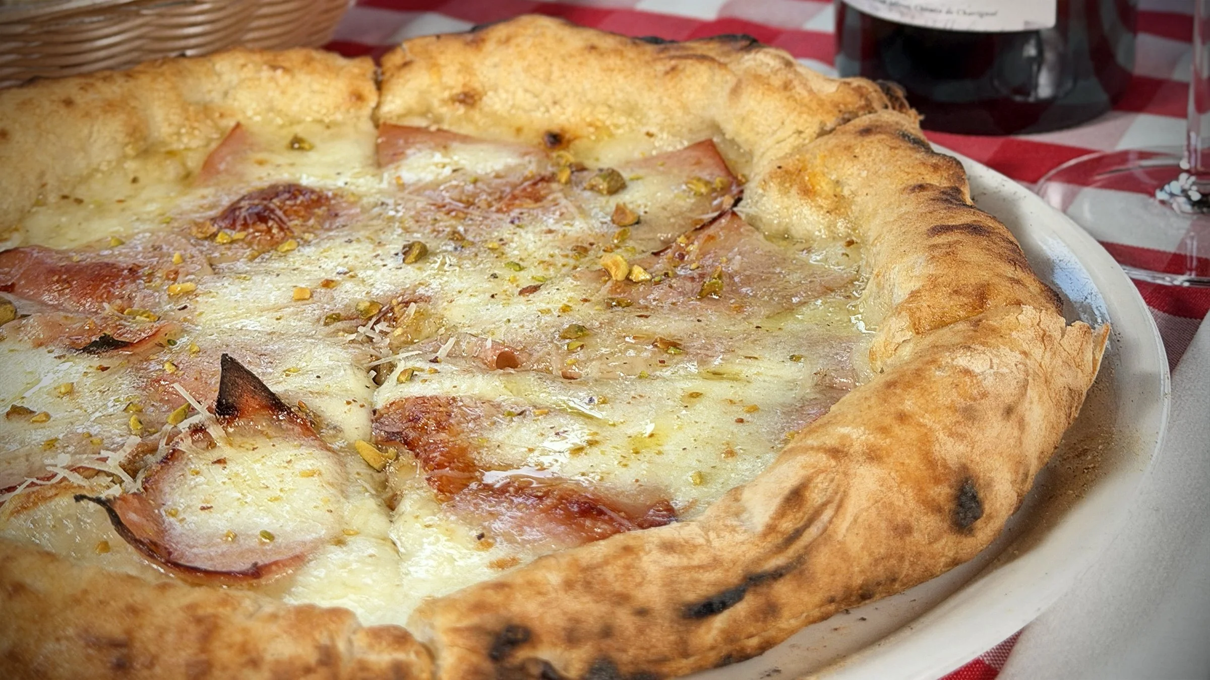 Close-up of a freshly baked pizza with cheese, ham, and sprinkled pistachios on top, placed on a white plate with a red and white checkered tablecloth in the background.