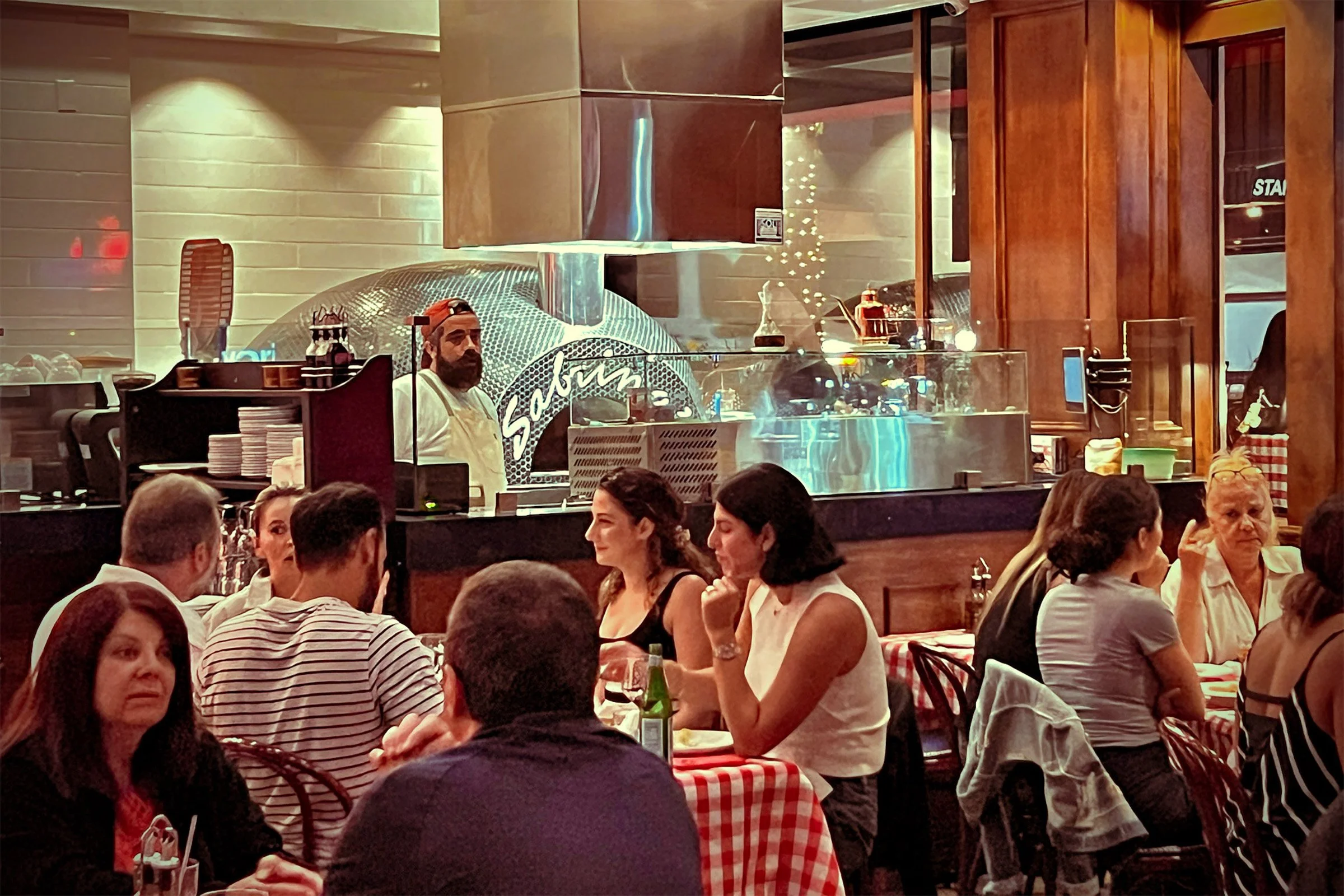 A busy restaurant with diners seated at tables covered with red and white checkered tablecloths, and a chef preparing food behind a glass partition with a pizza oven in the background.