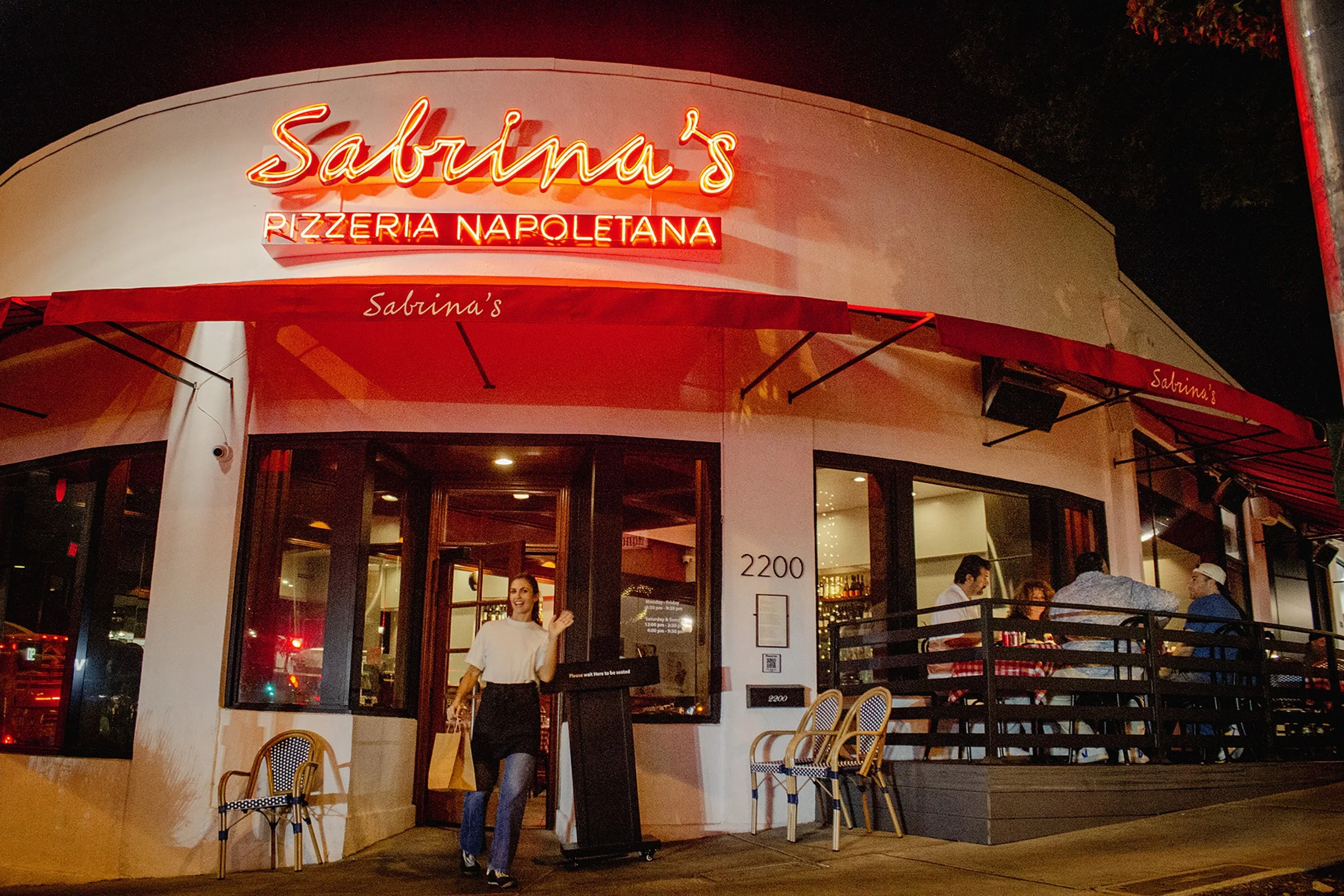 Night view of Sabrina's Pizzeria Napoletana storefront with red neon sign, woman exiting, and patrons on the outdoor patio